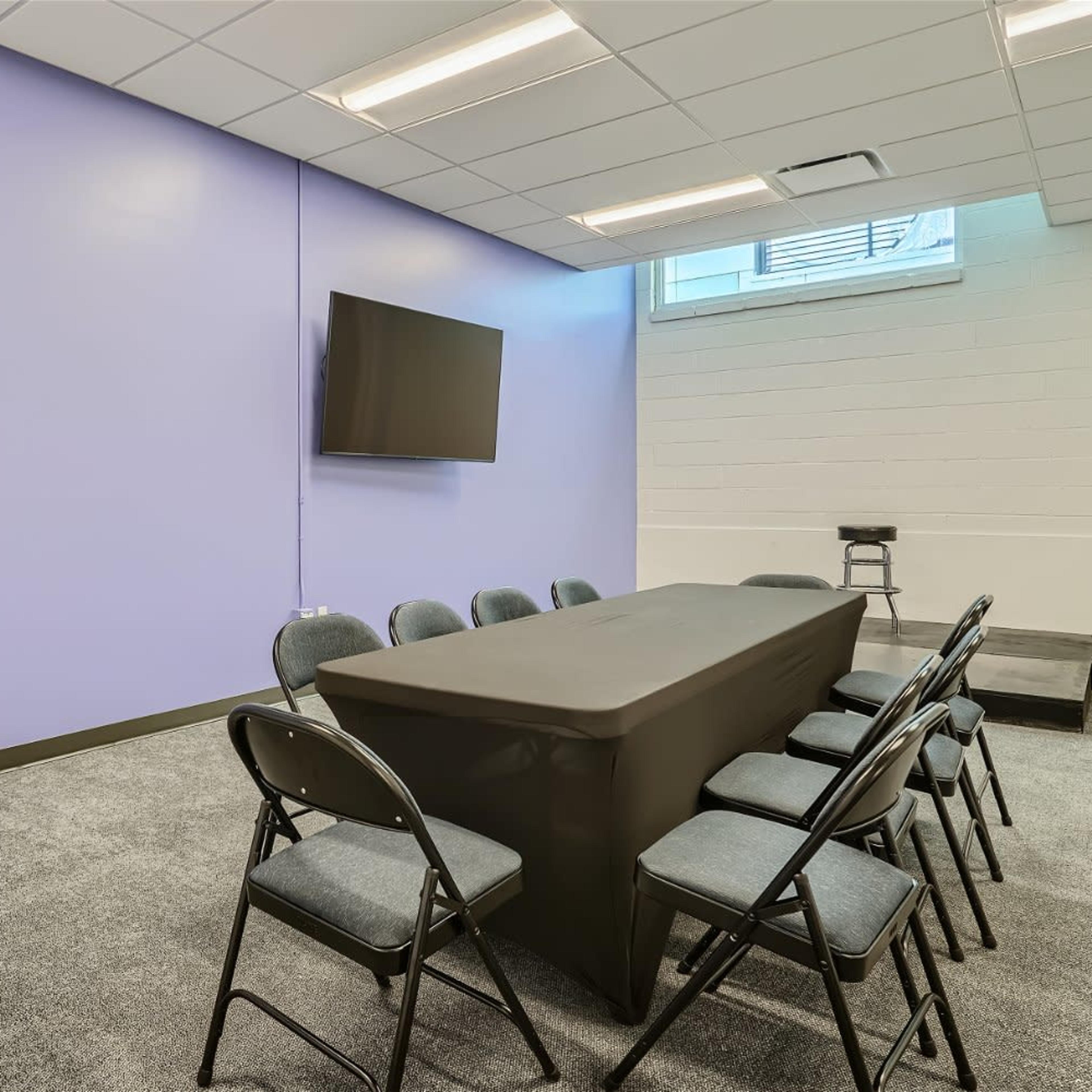 A conference room with a long black table surrounded by eight chairs, a television mounted on the wall, and a light purple wall color.