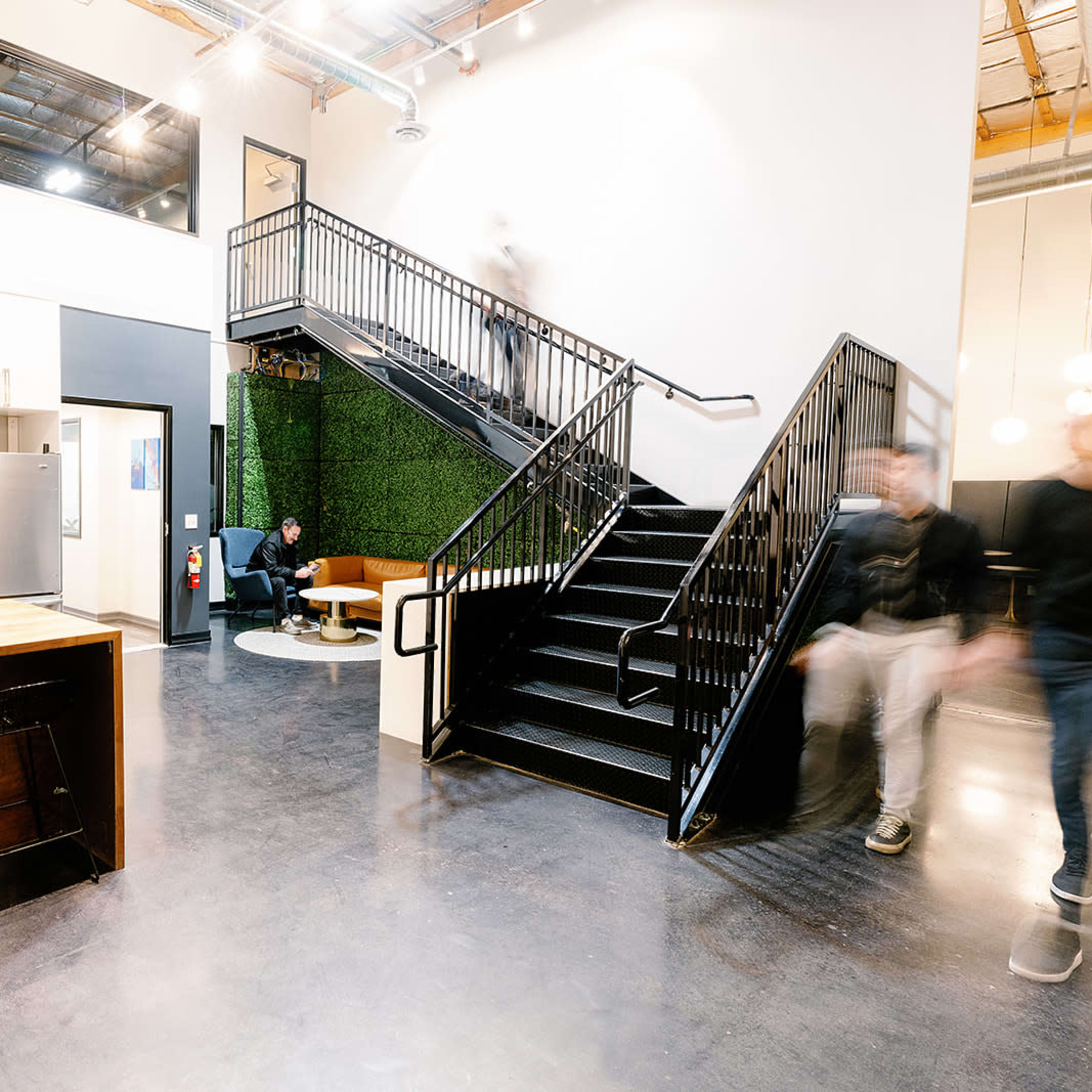 A modern open-plan workspace featuring a kitchen area with bar stools, a green wall, and a staircase leading to an upper level.