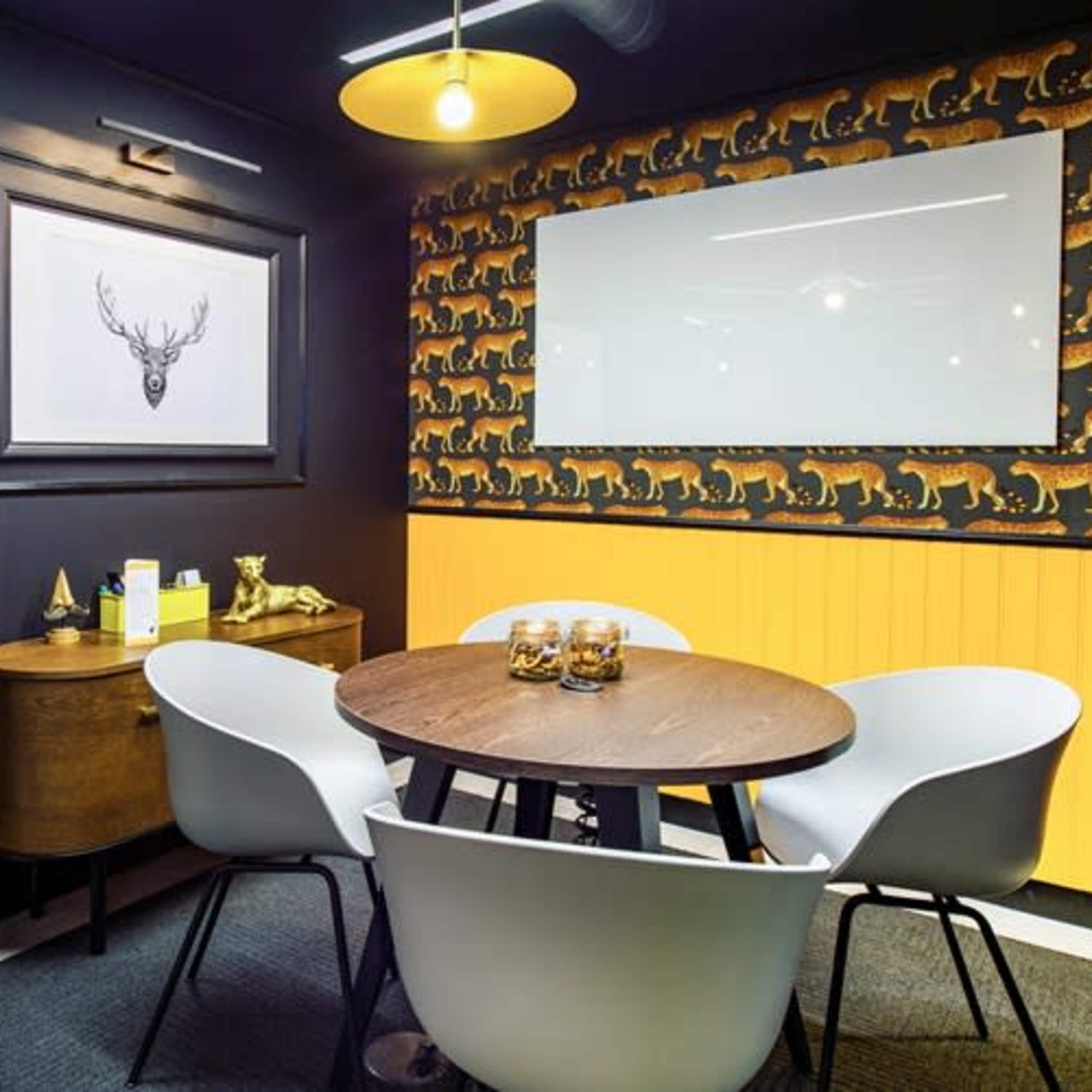 A small meeting room features a round wooden table surrounded by white chairs, a deer print on the wall, and a patterned wallpaper depicting animals.