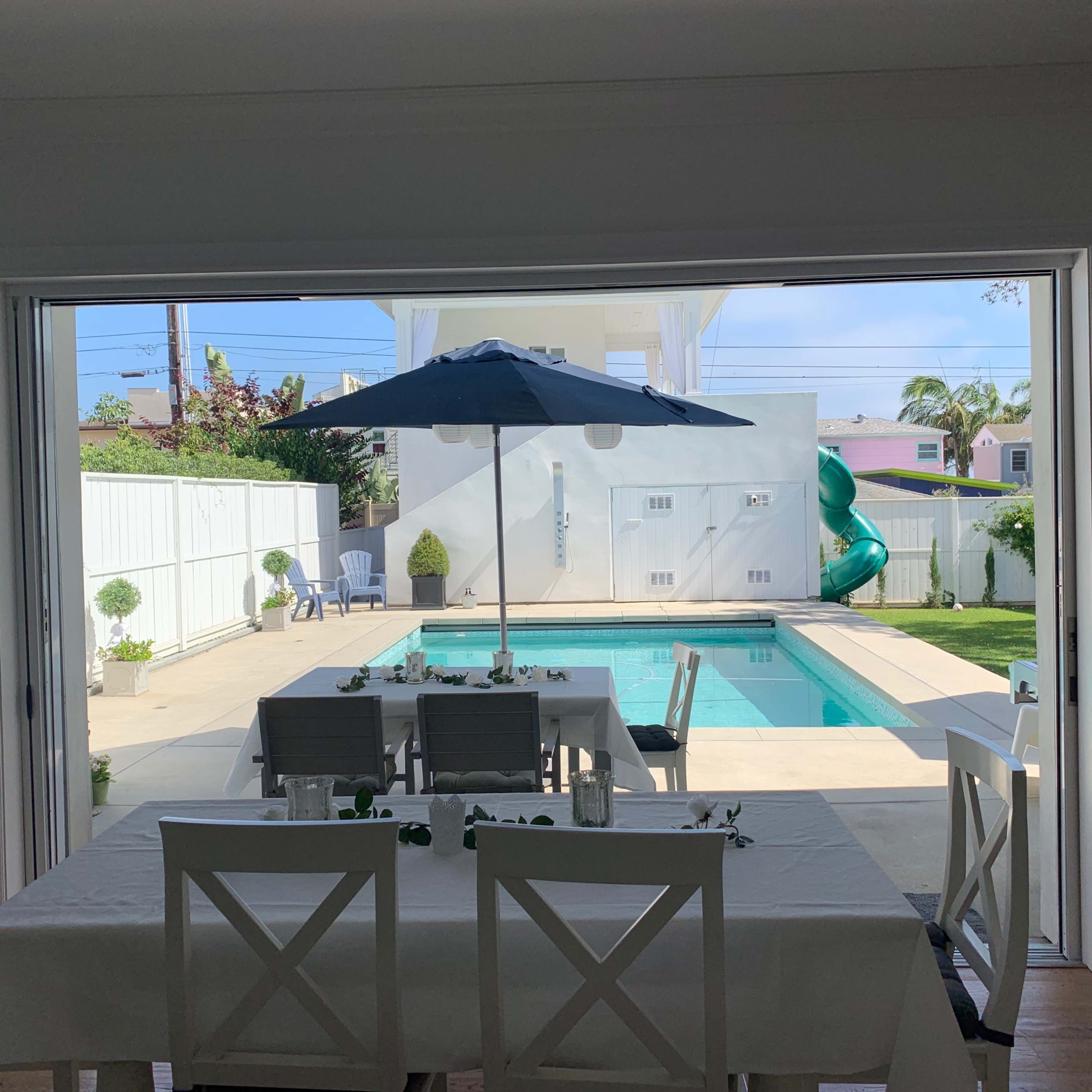 A dining area with a table under an umbrella overlooks a swimming pool and a backyard.