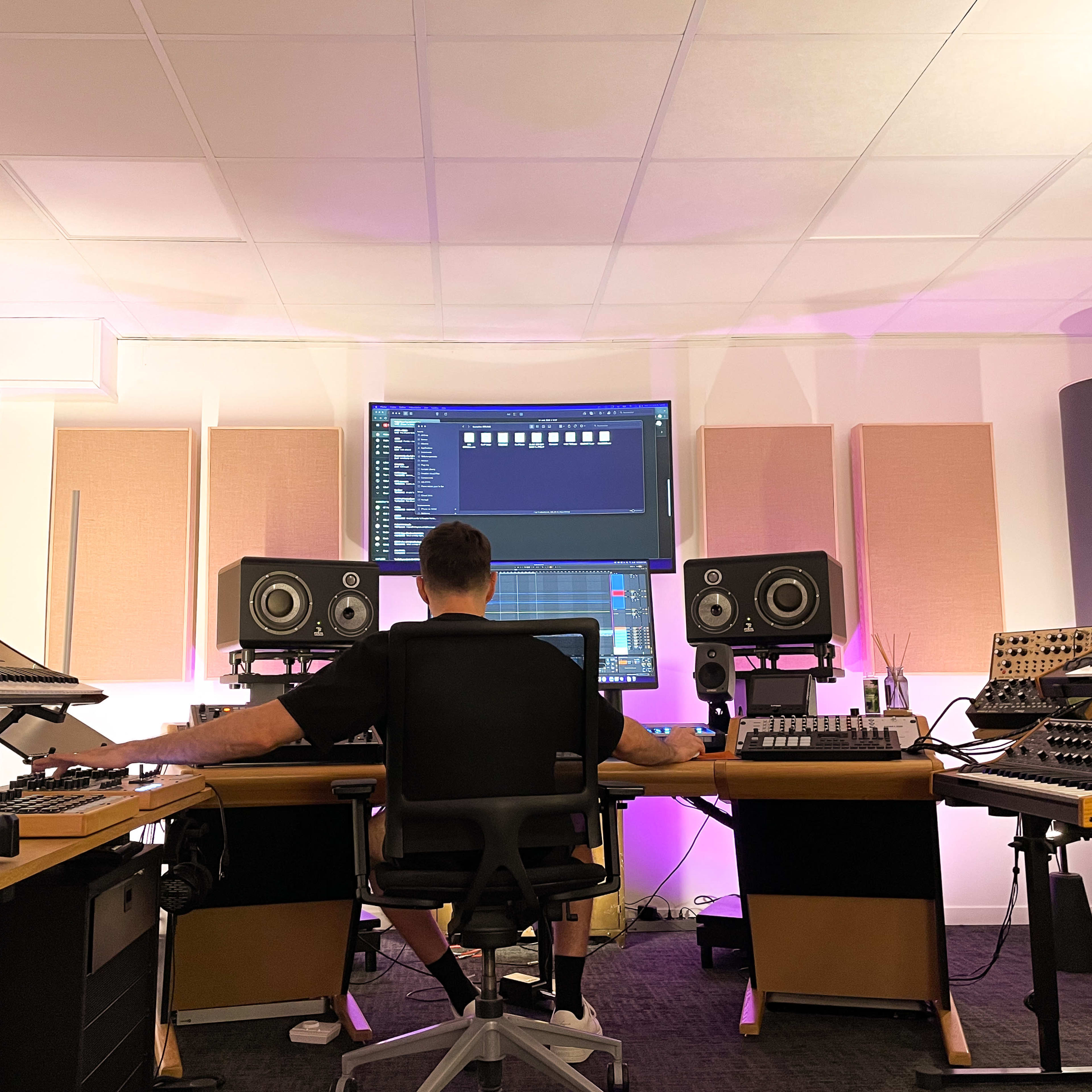 A person is seated at a mixing console in a music studio, surrounded by various sound equipment and displays.