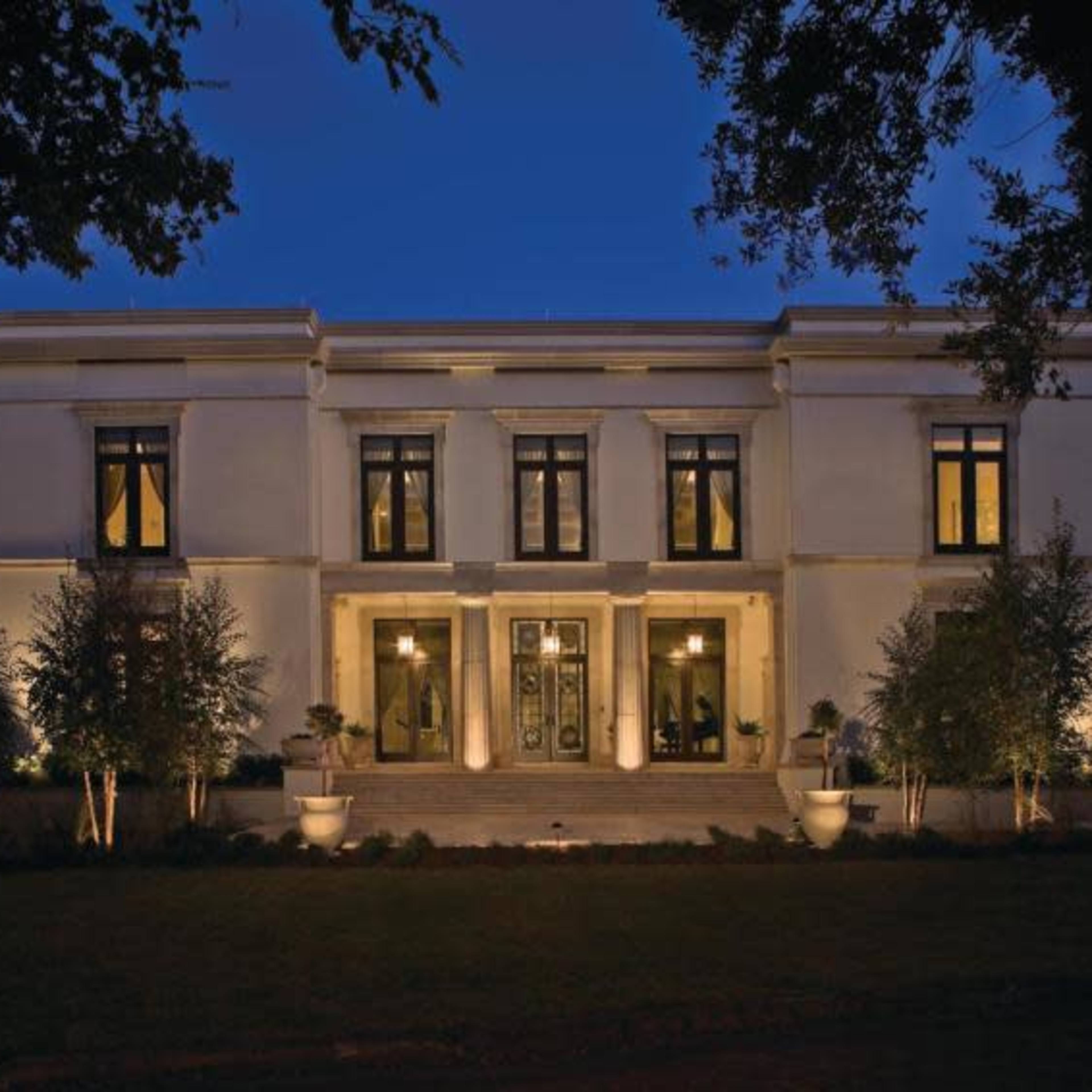 A modern, two-story white house with large windows and symmetrical features is illuminated by outdoor lighting at twilight.