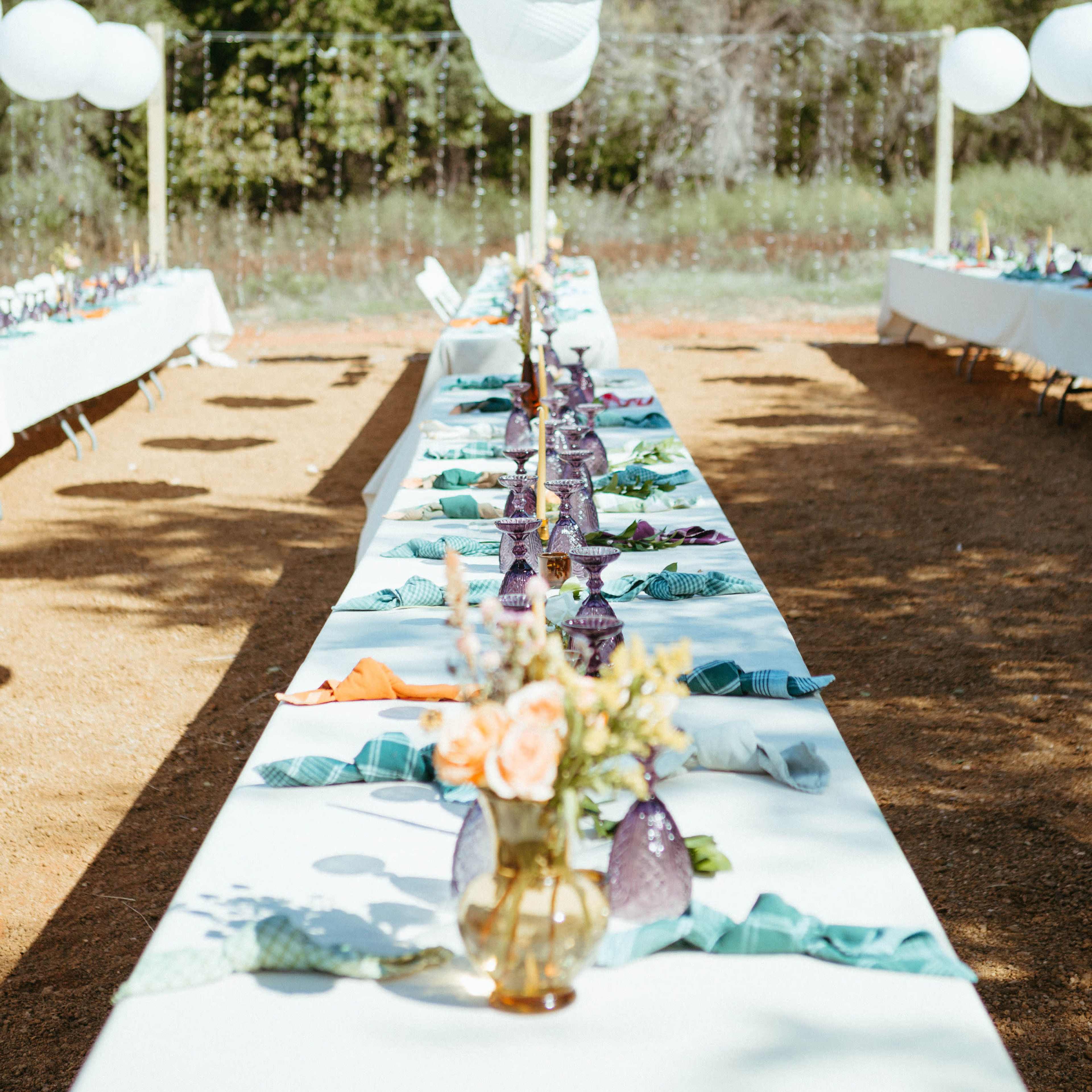 A long dining table is set outdoors, adorned with white tablecloths, colorful napkins, and vases of flowers, while oversized white lanterns hang above.