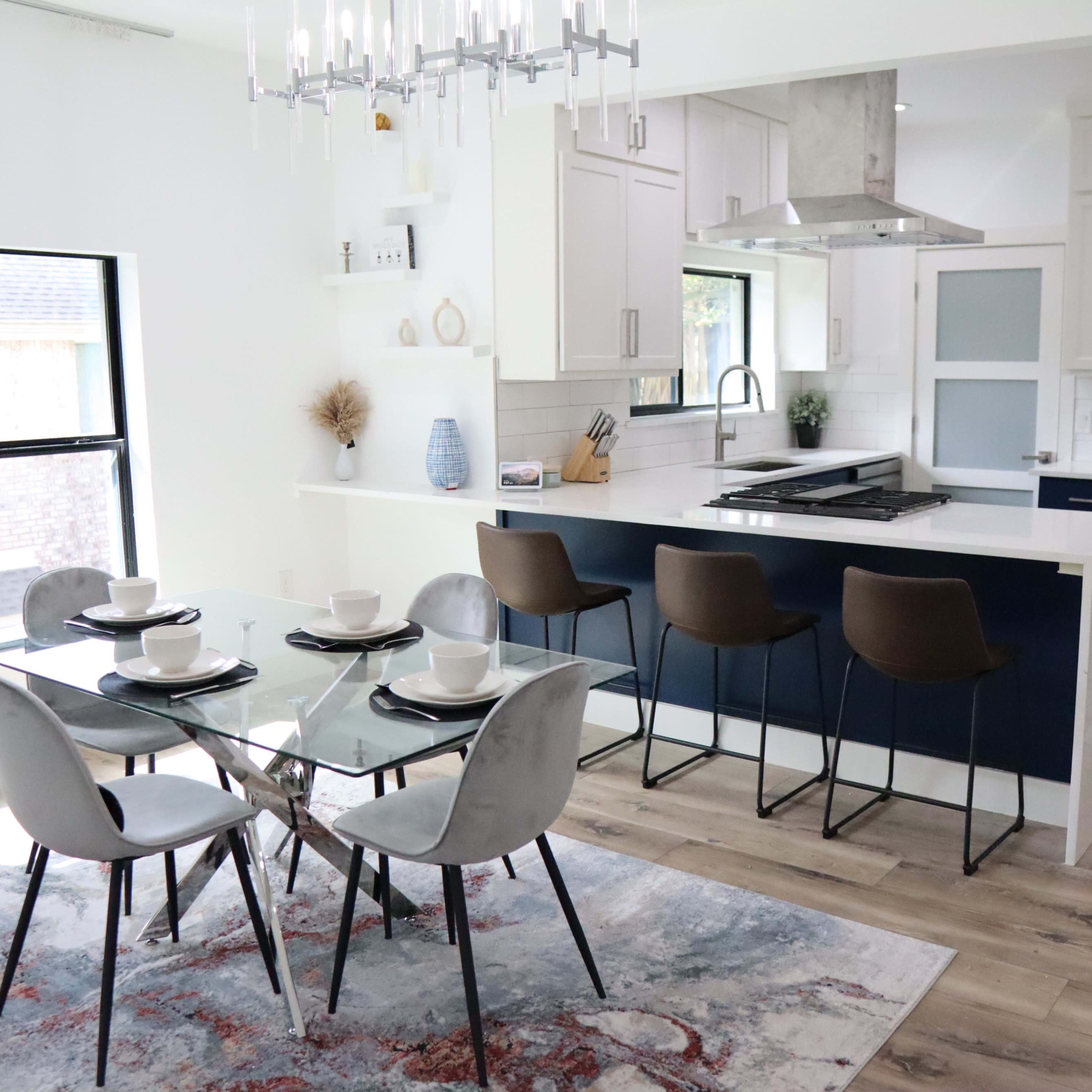 A modern kitchen and dining area feature a glass dining table with four chairs, a kitchen island with bar stools, and white cabinetry with navy blue accents.