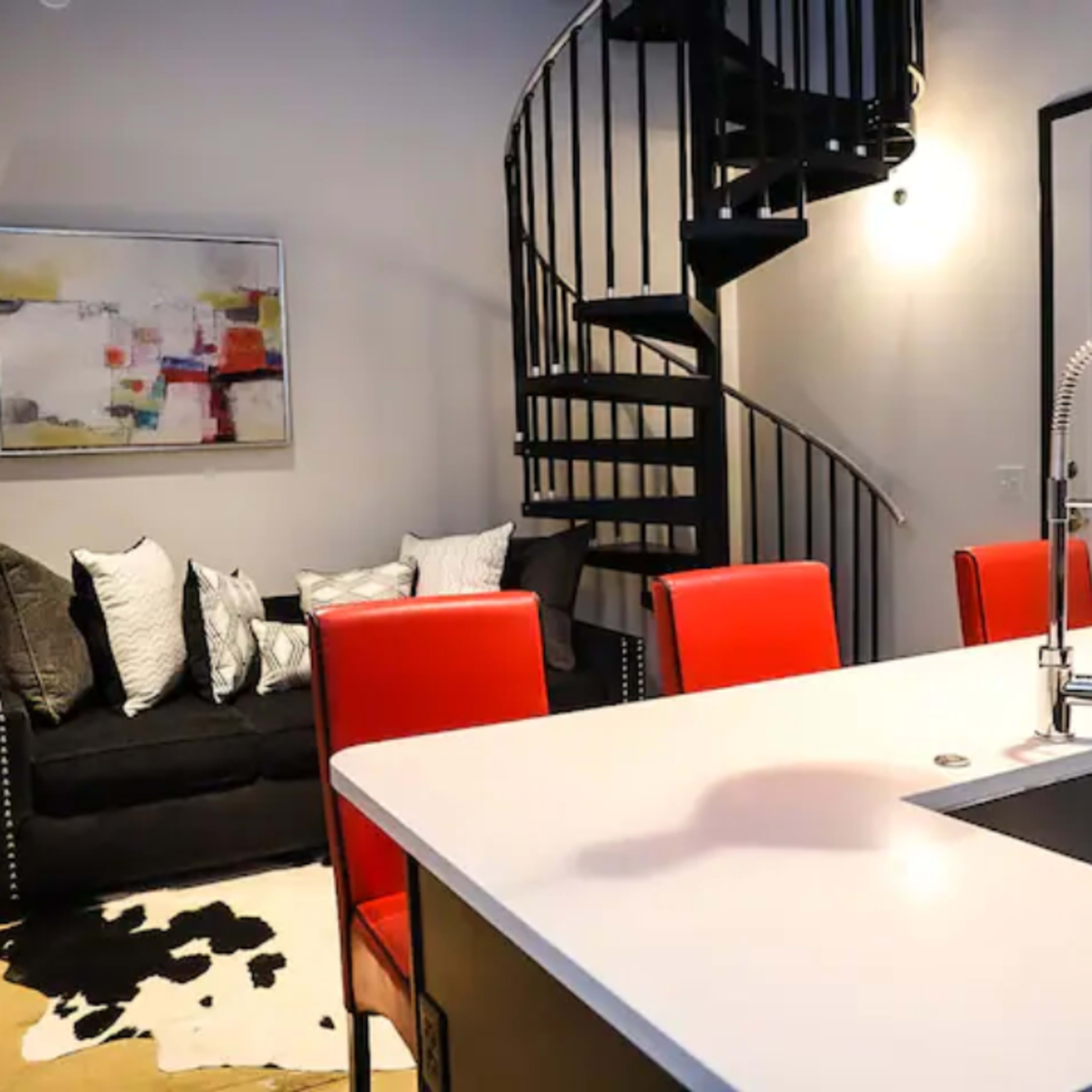 A modern living space featuring a spiral staircase, a black sofa with decorative pillows, a white kitchen island with red chairs, and abstract wall art.