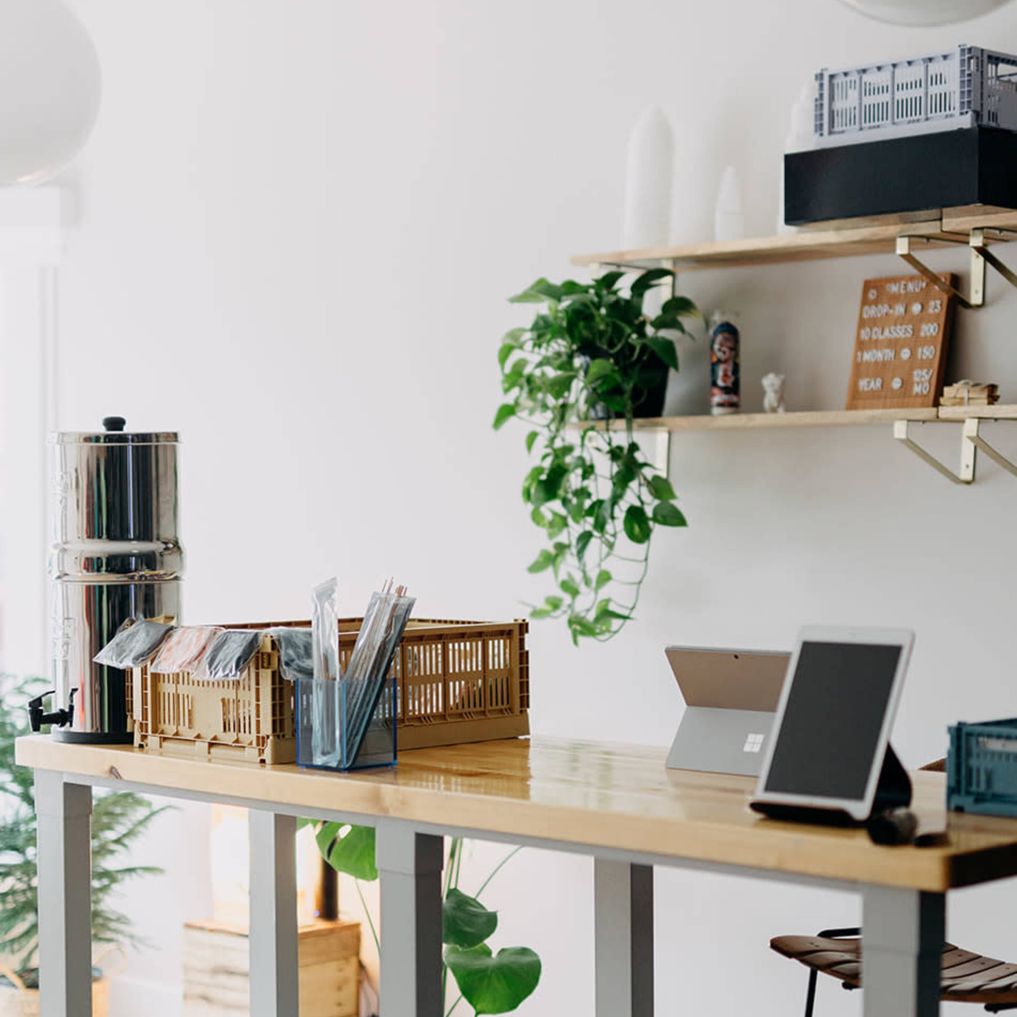 A modern workspace features a long wooden table with a beverage dispenser and organizational baskets, against a backdrop of greenery and minimalist decor.