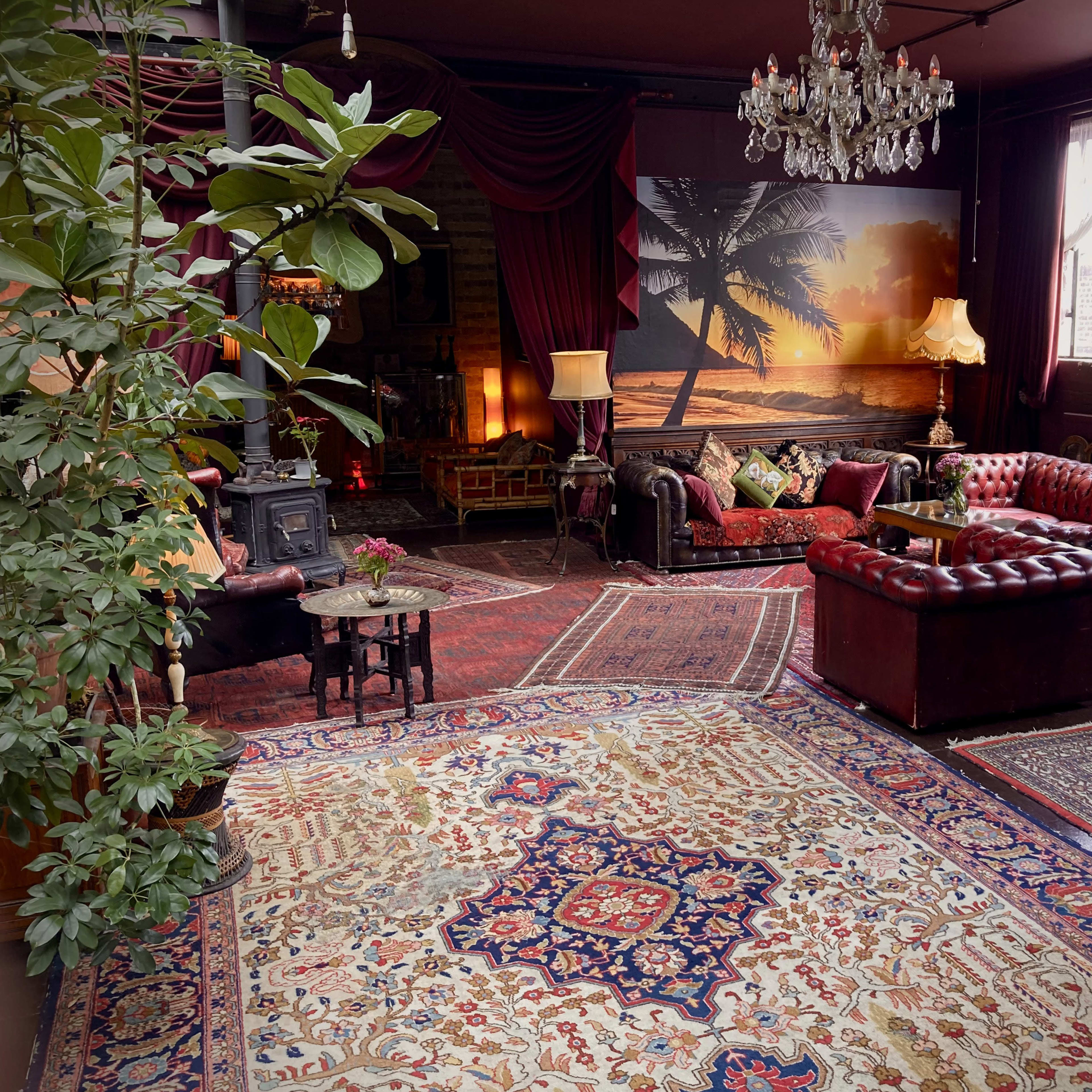 An elegantly furnished living room features leather sofas, a large decorative rug, a chandelier, and a sunset mural with palm trees.