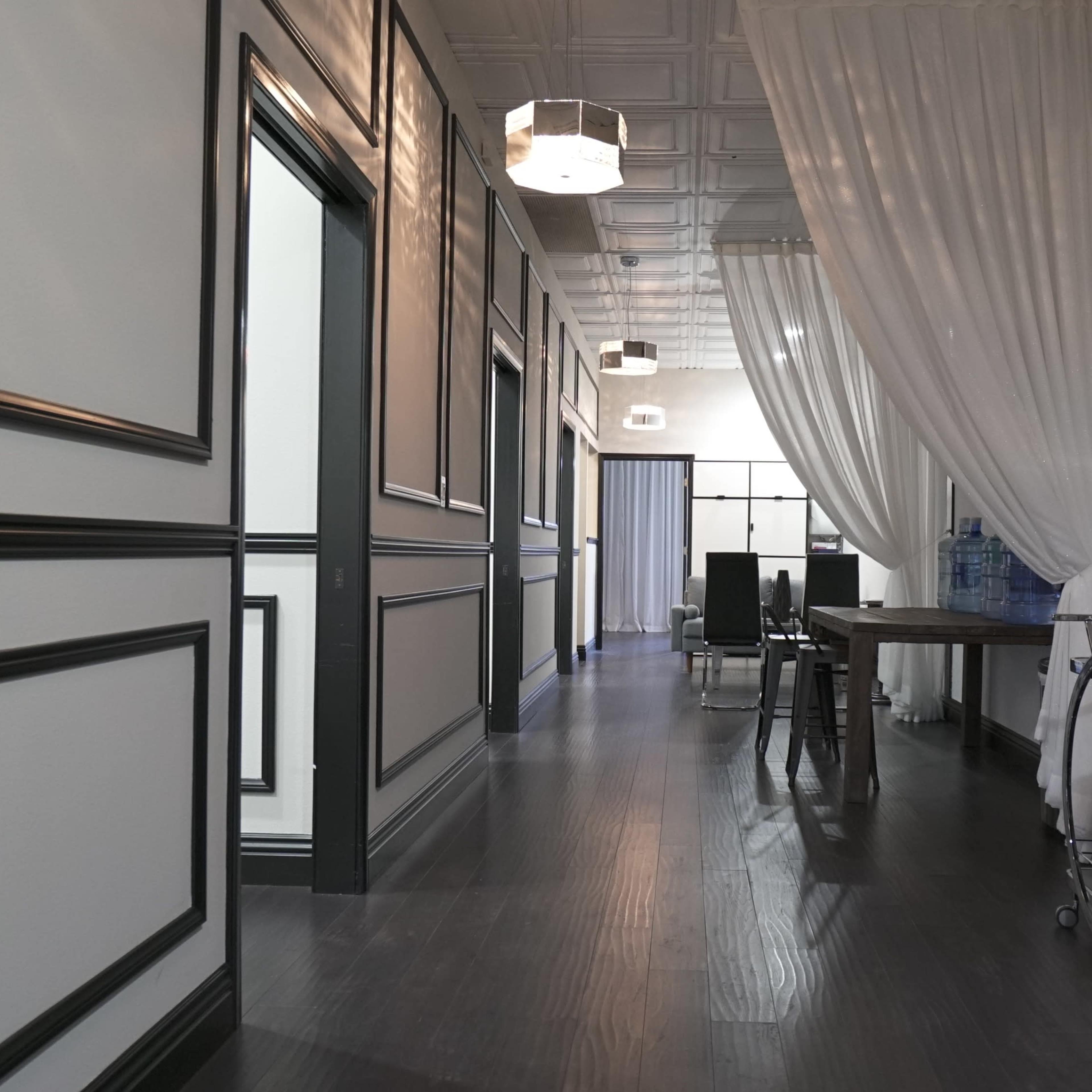 A hallway with a dark wooden floor, framed walls, and decorative lighting fixtures, leading to a seating area and a glass water dispenser.