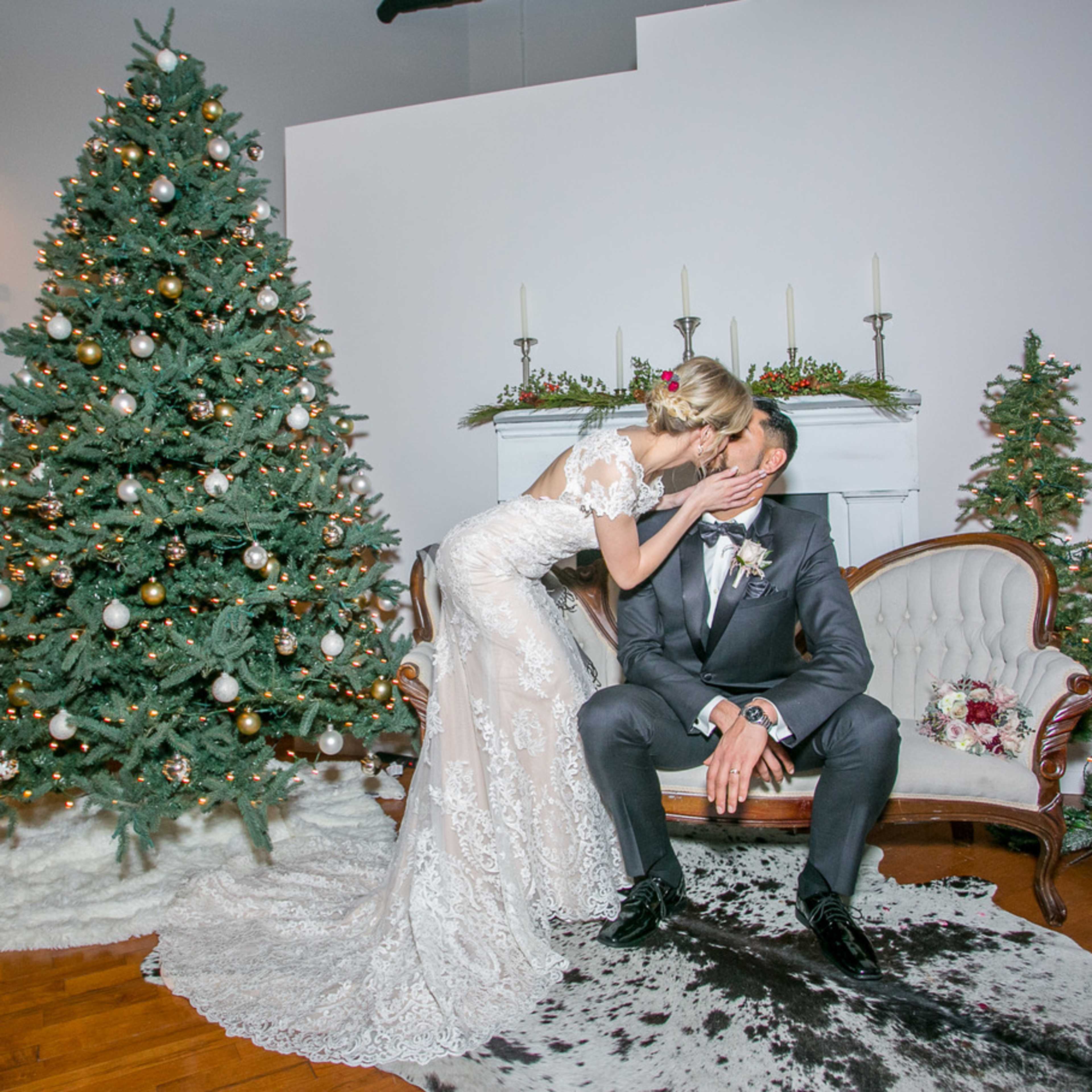 A bride kisses a groom while sitting on a vintage sofa, with a decorated Christmas tree and smaller evergreen trees in the background.