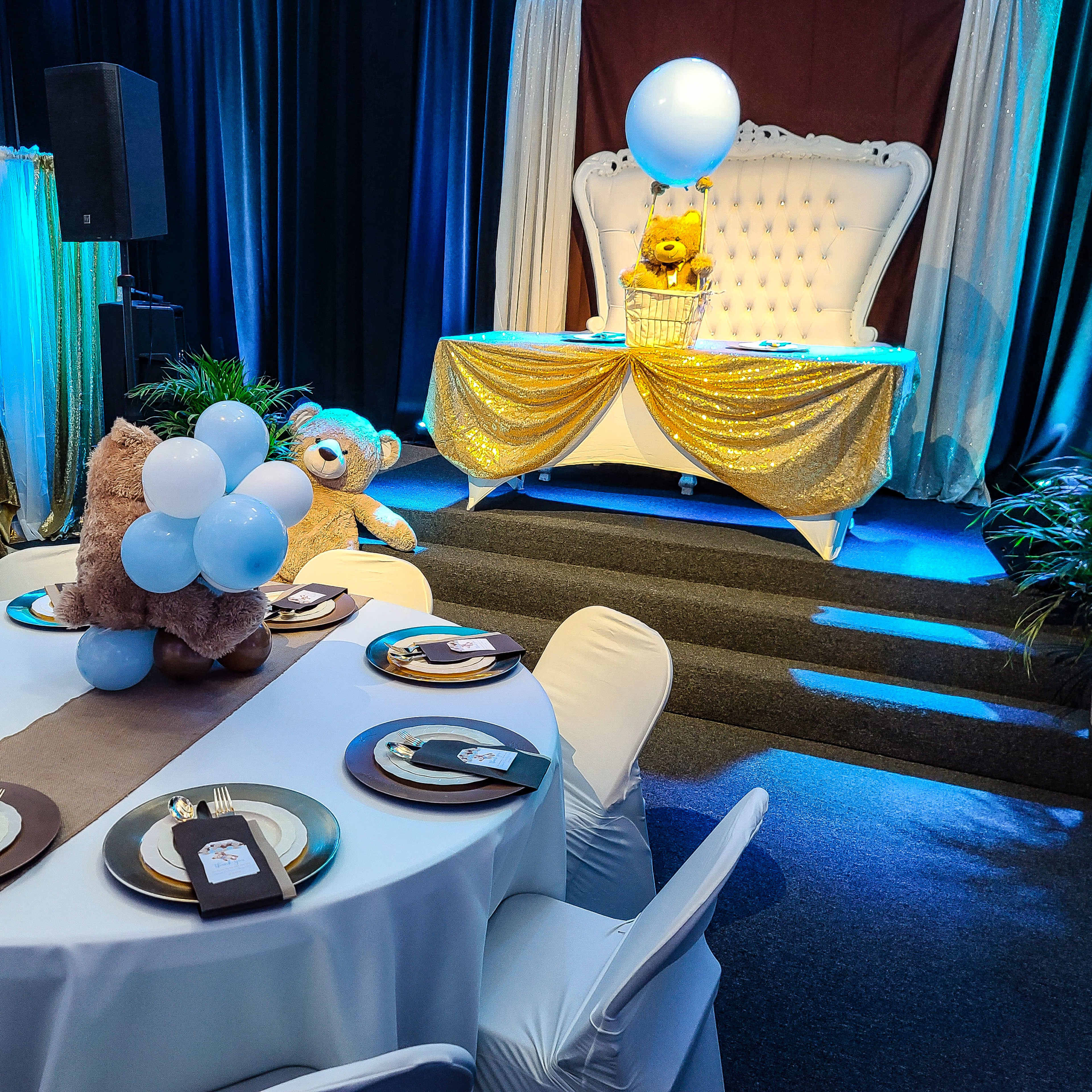 The scene shows a decorated event space with a table set for a celebration, featuring white tablecloths, golden accents, and a backdrop of a plush throne with teddy bears and balloons.