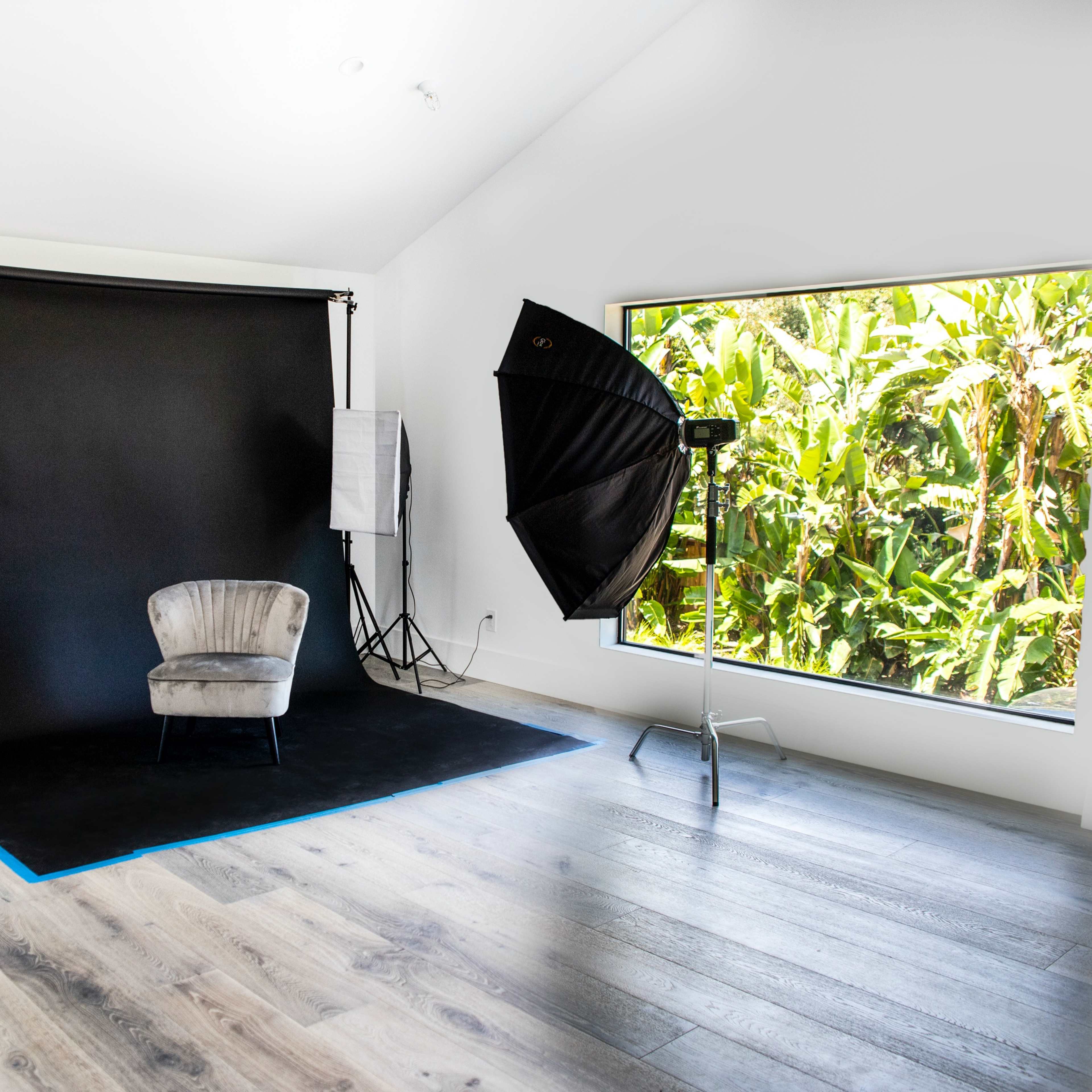 A photography studio features a gray armchair positioned on a black backdrop with large windows revealing lush greenery outside.