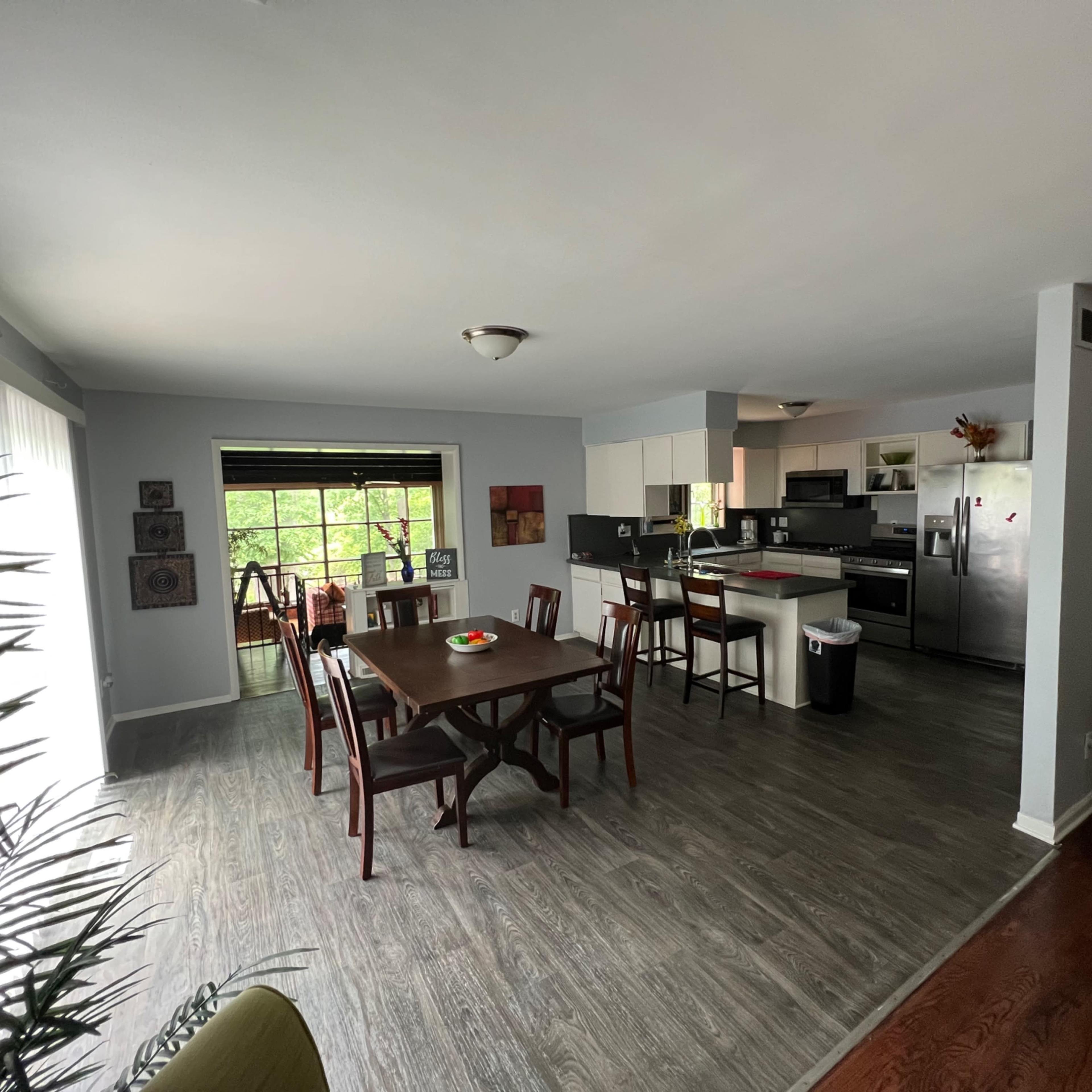A spacious dining area with a wooden table and chairs next to a kitchen, featuring light gray walls and large windows.