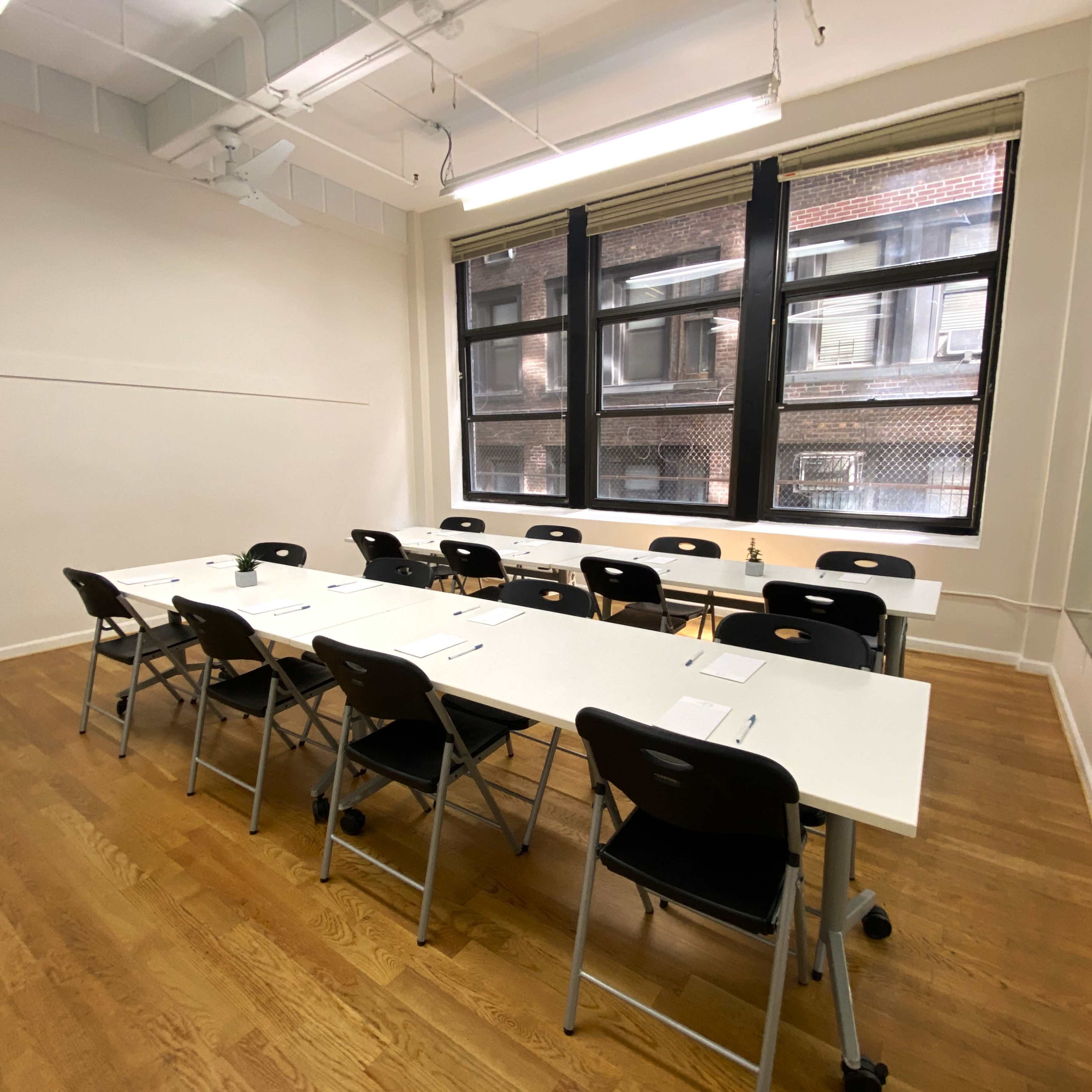 A classroom features a long row of white tables and black chairs arranged neatly, along with large windows allowing natural light to fill the space.
