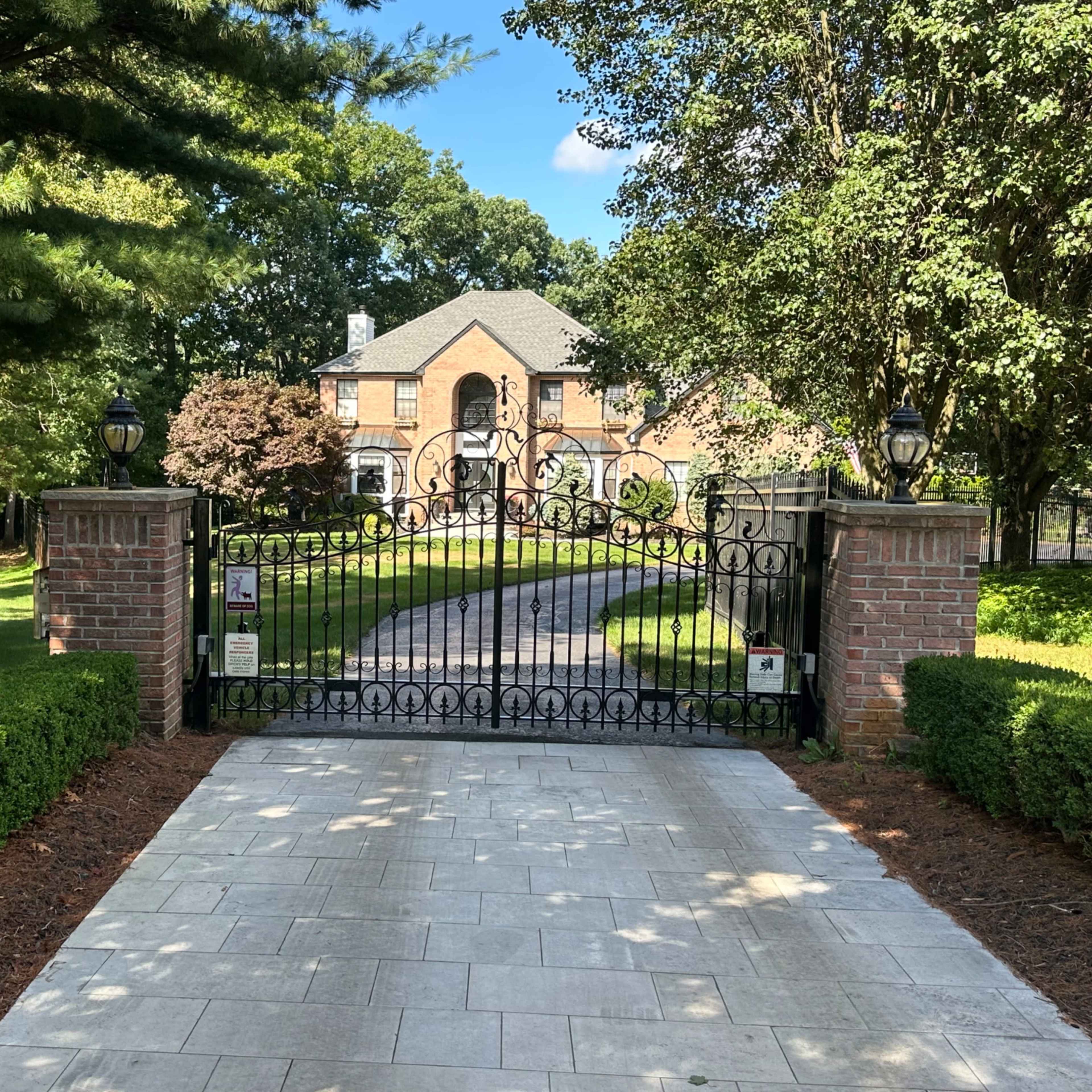 A wrought iron gate leads to a large brick house surrounded by greenery and trees.
