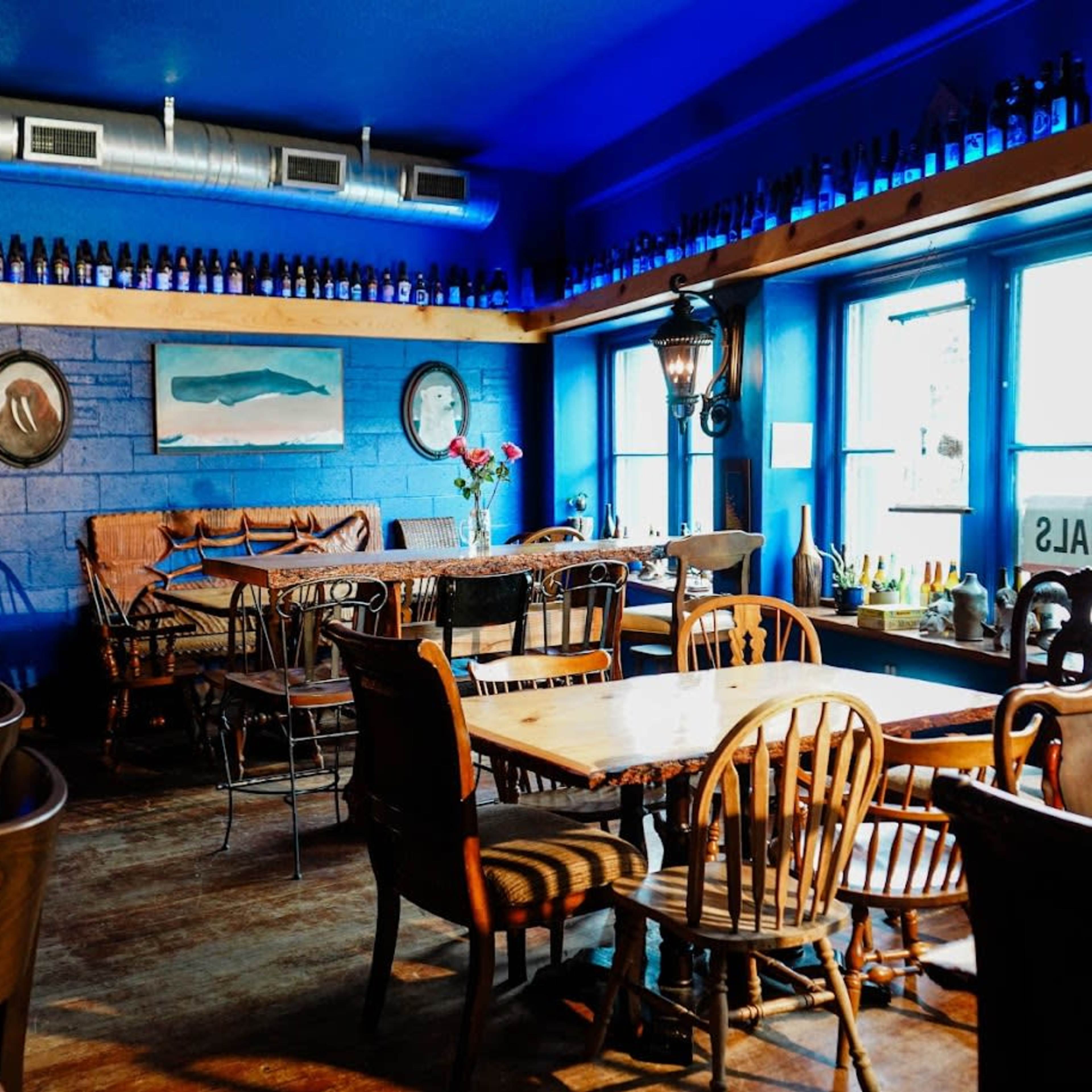 A cozy restaurant interior featuring blue walls, wooden tables, and various bottles displayed on a shelf above the windows.