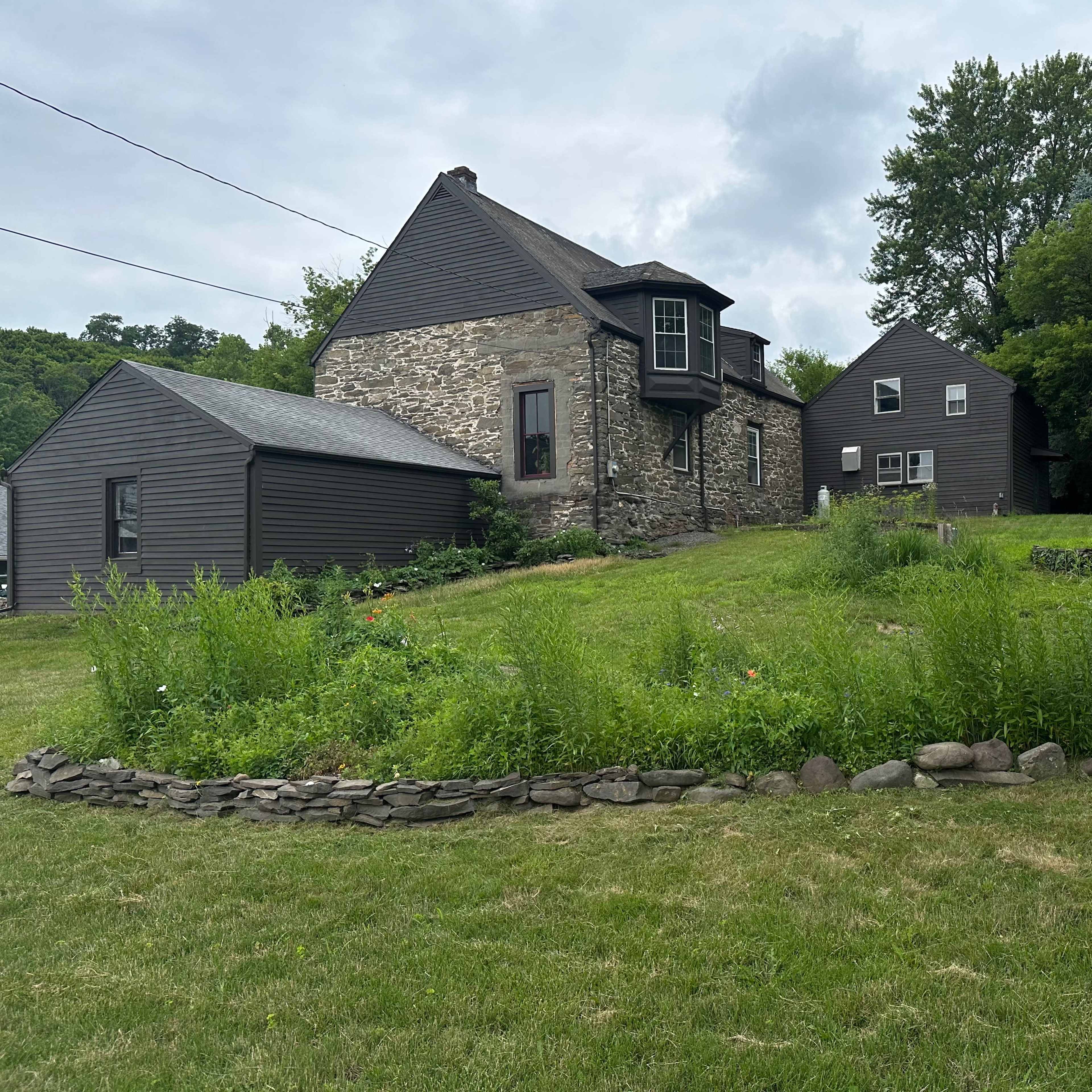 A grassy area featuring two houses, one made of stone and the other clad in dark siding, surrounded by trees and a stone-bordered garden.
