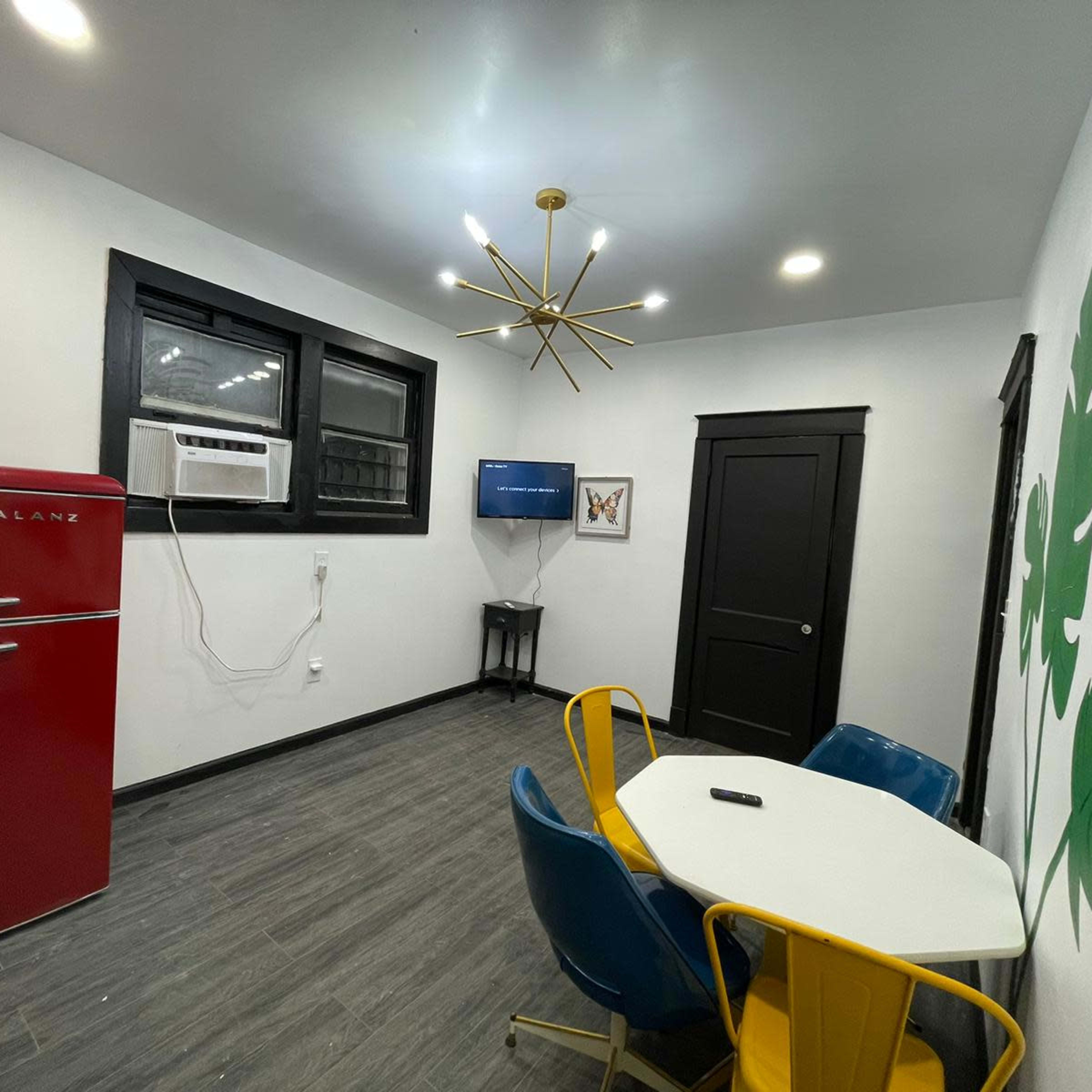 A modern kitchen area with a red retro-style refrigerator, an air conditioning unit, a small round table surrounded by blue and yellow chairs, and a wall with a green leaf design.