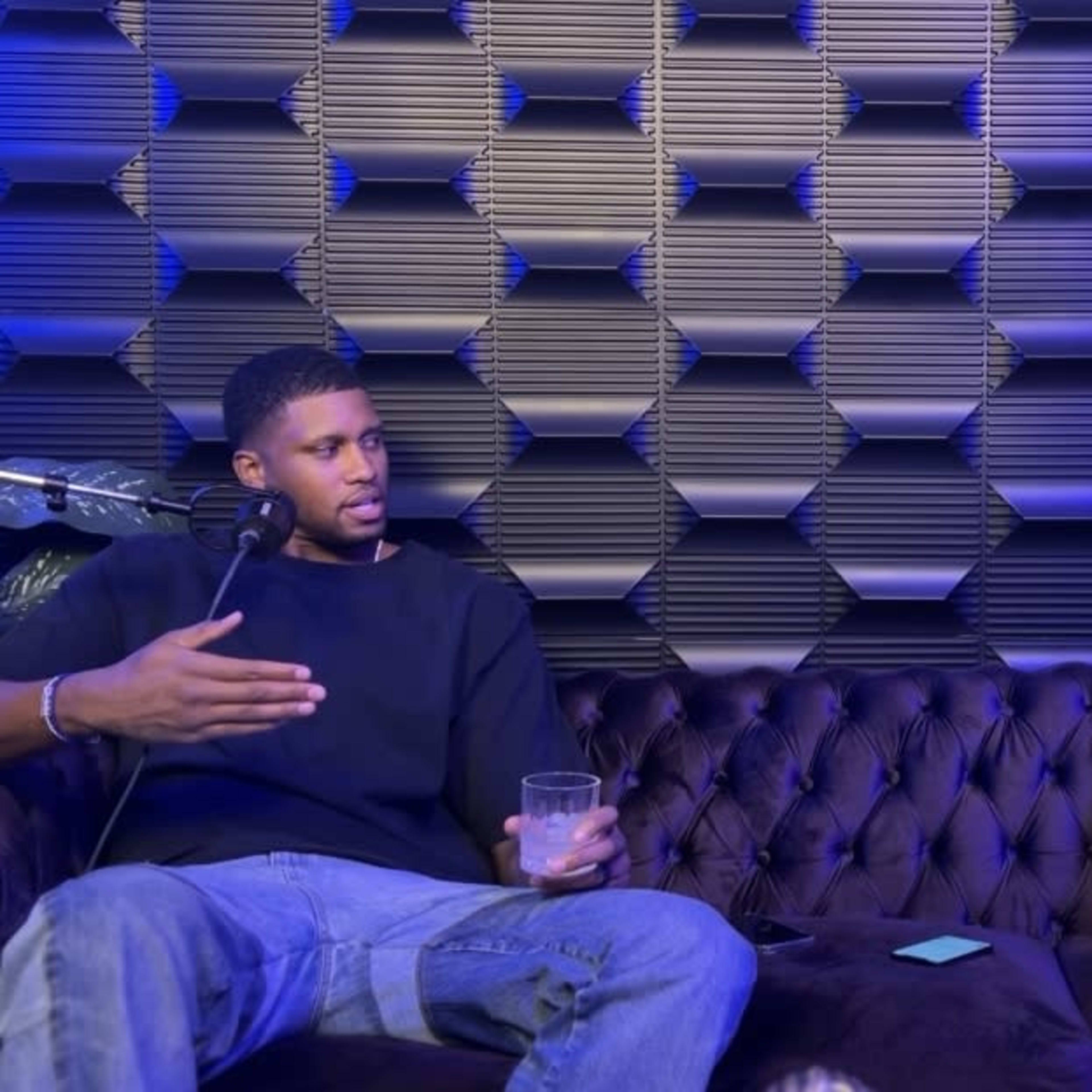 Two men are seated on a couch in front of microphones, engaged in conversation against a textured blue wall.