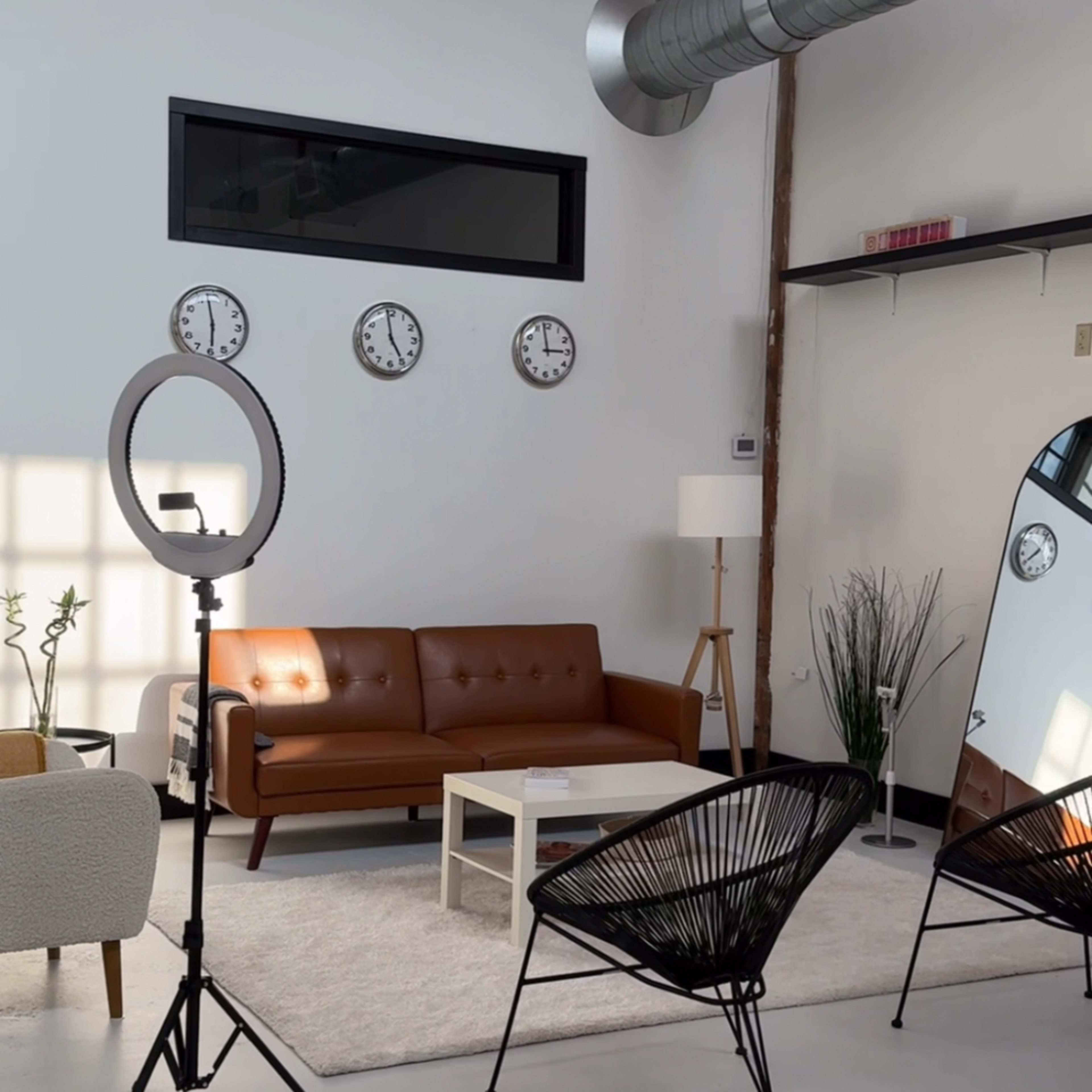 A modern interior space featuring a brown leather sofa, a white coffee table, three wall clocks, and two black chairs beside a large mirror.