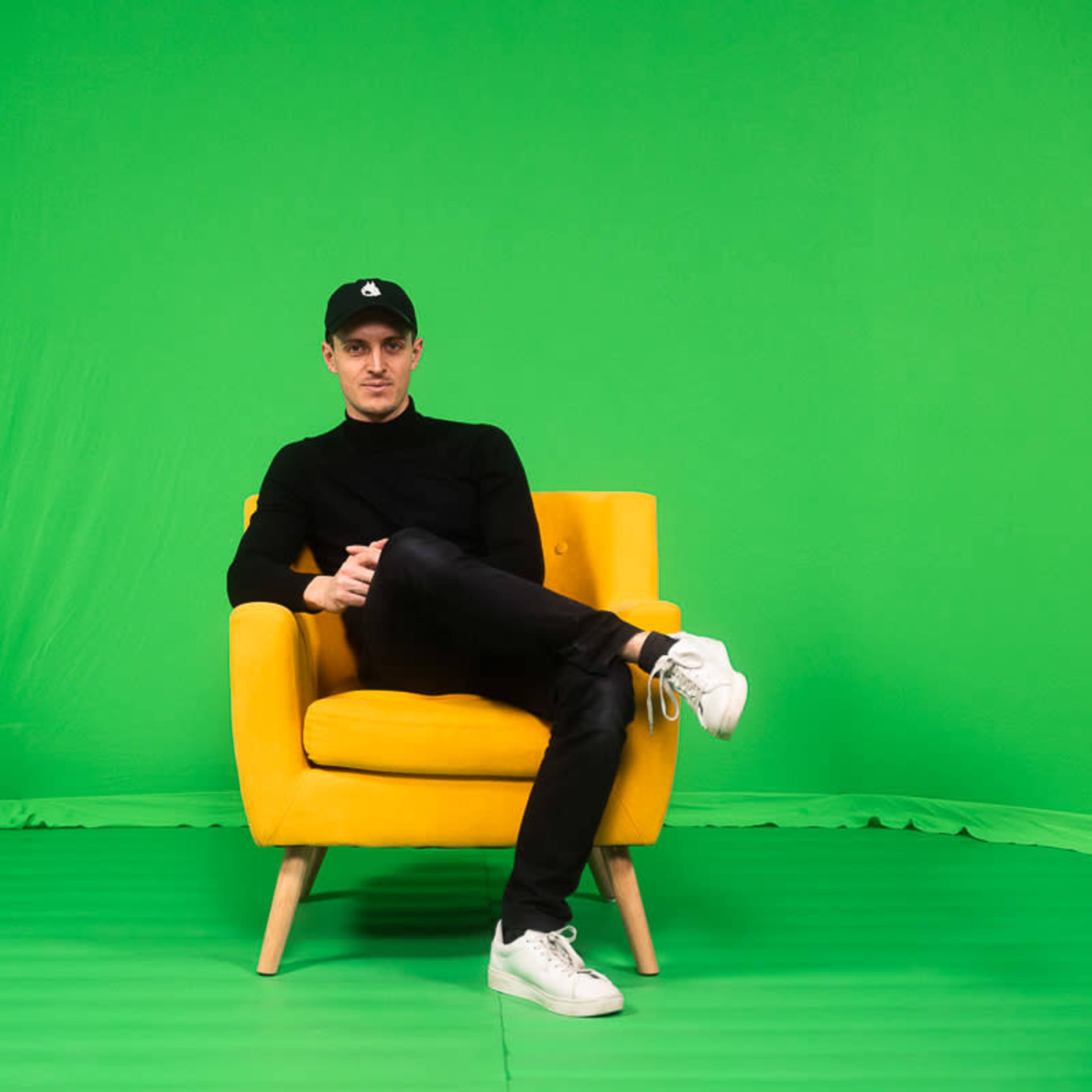 A person sits cross-legged in a yellow chair against a green backdrop.