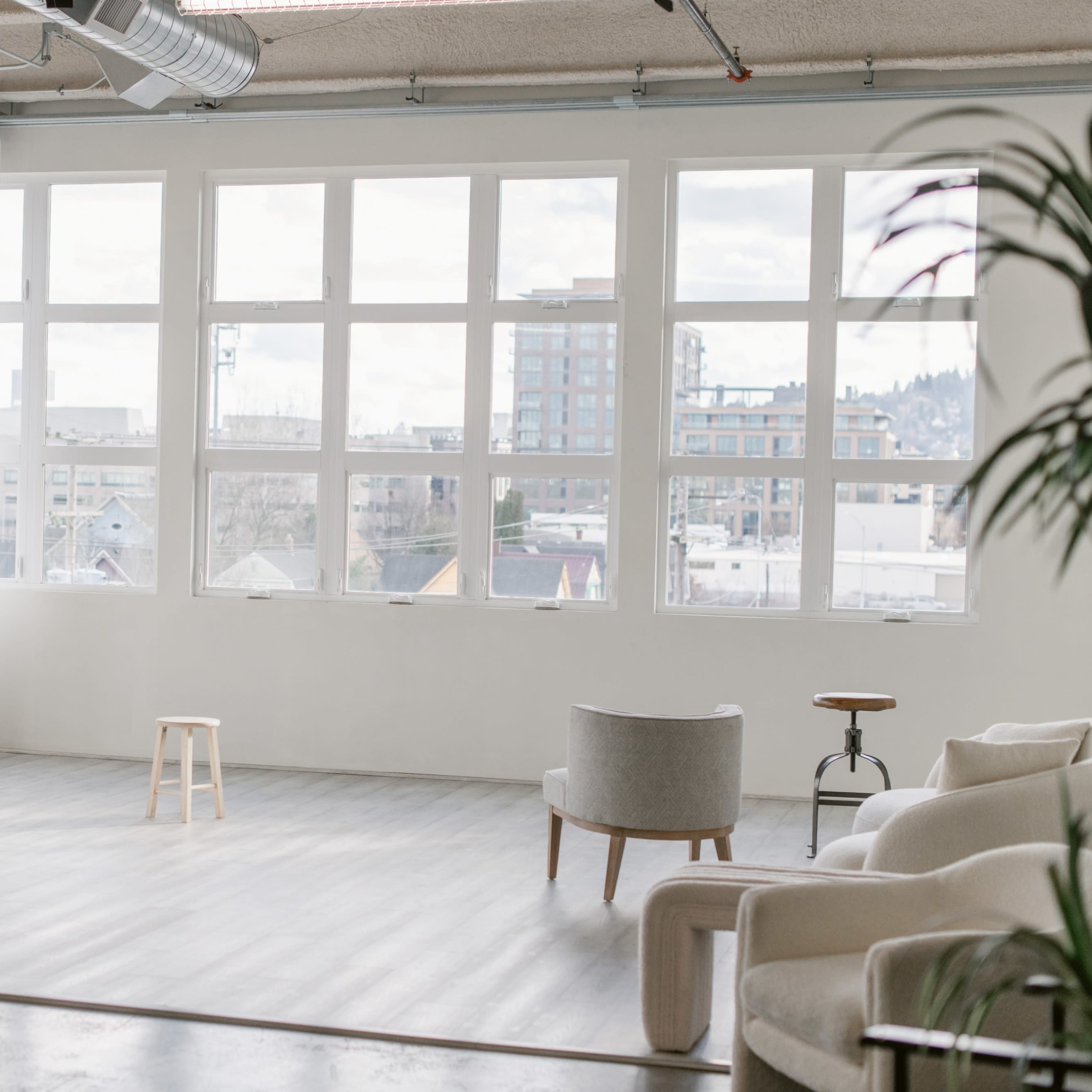 A spacious, well-lit room with large windows, a stool in the center, and a chair against the wall.