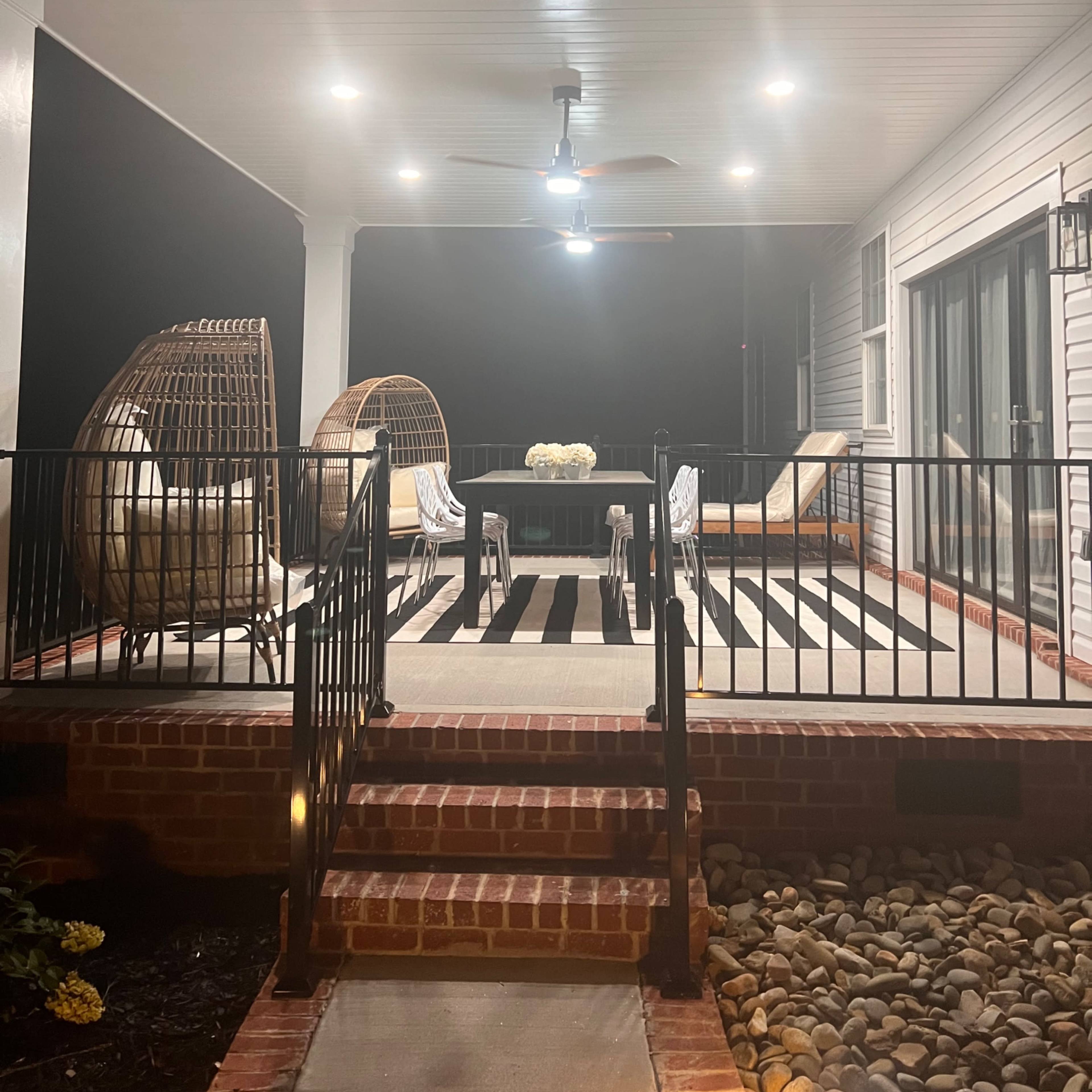 A covered outdoor patio with two wicker chairs, a table with a floral centerpiece, and a ceiling fan, illuminated by overhead lights on a dark night.