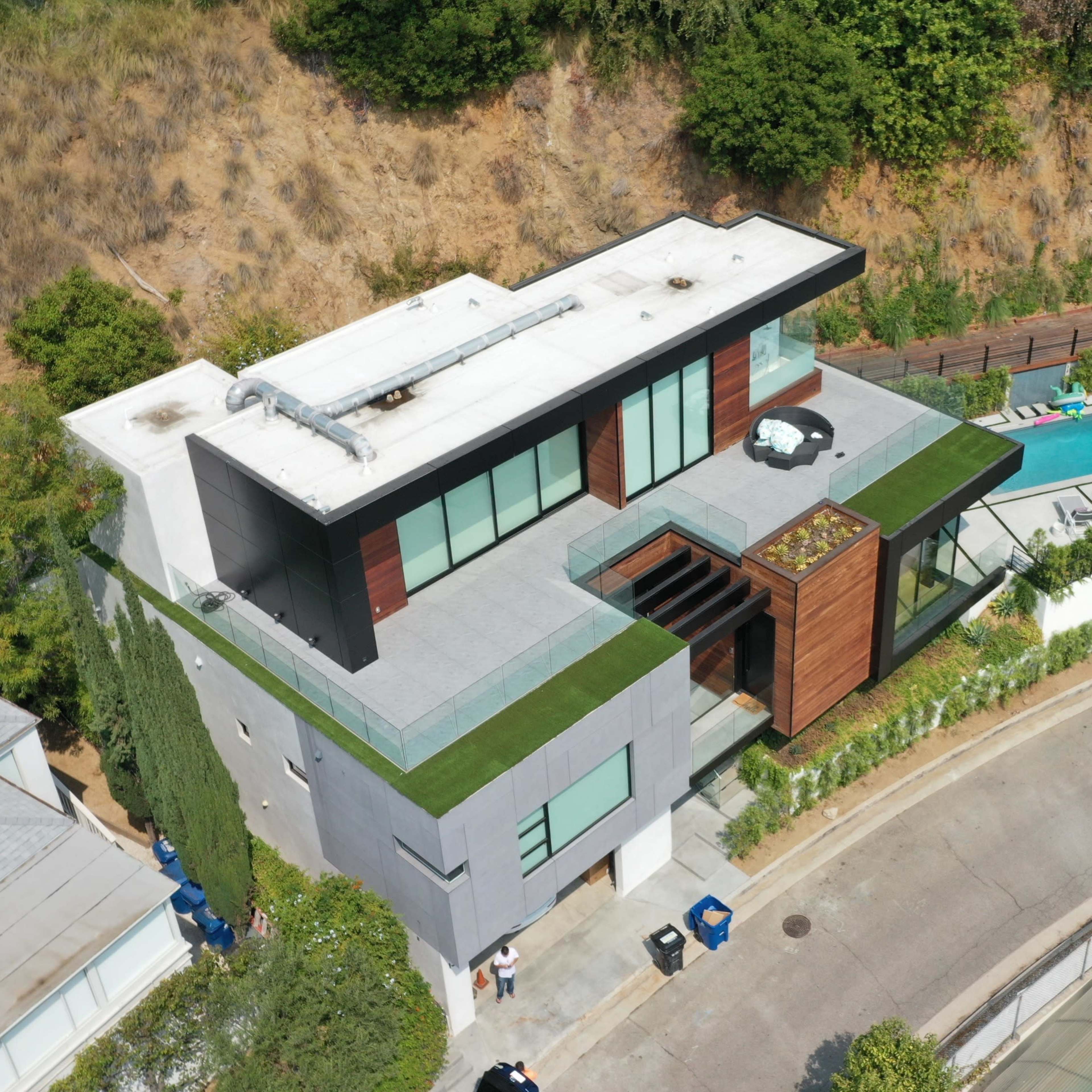 A modern house with a flat roof and green landscaping, situated on a hillside near a swimming pool and a winding road.