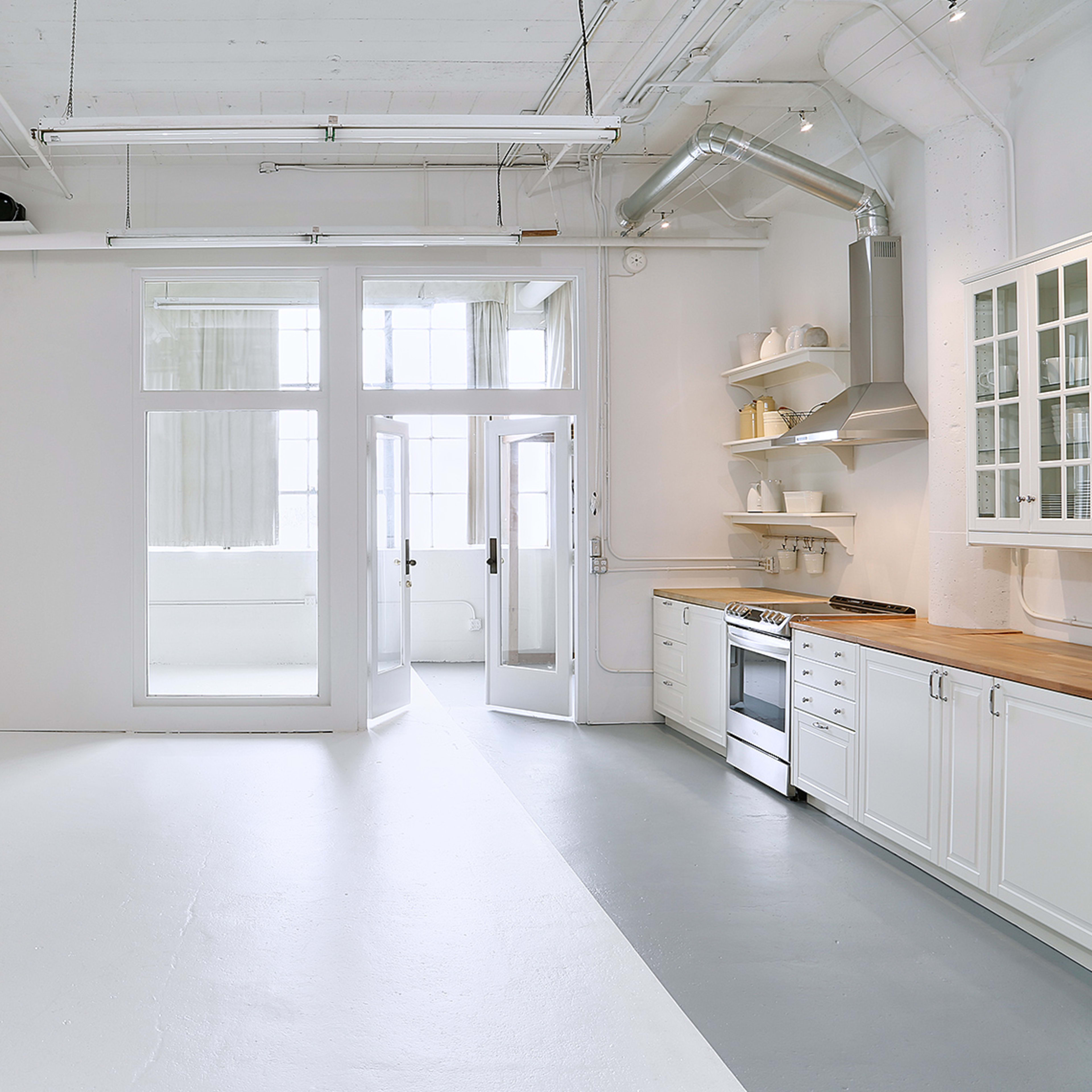 A modern, open kitchen with white walls, a wooden countertop, and large windows allowing natural light to enter.