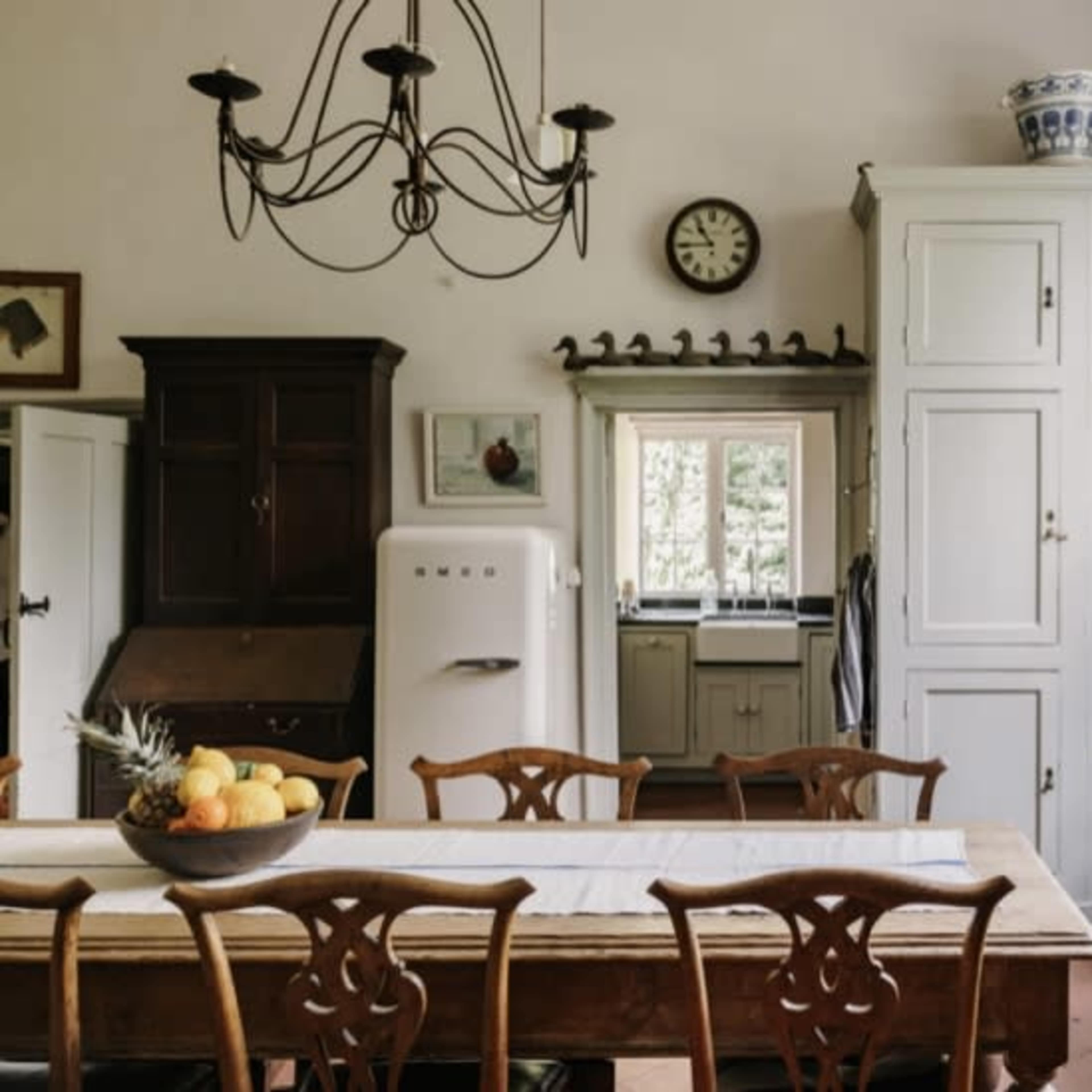 A well-lit kitchen features a large wooden table, a vintage refrigerator, and a white cabinet, with windows providing a view of the outdoors.