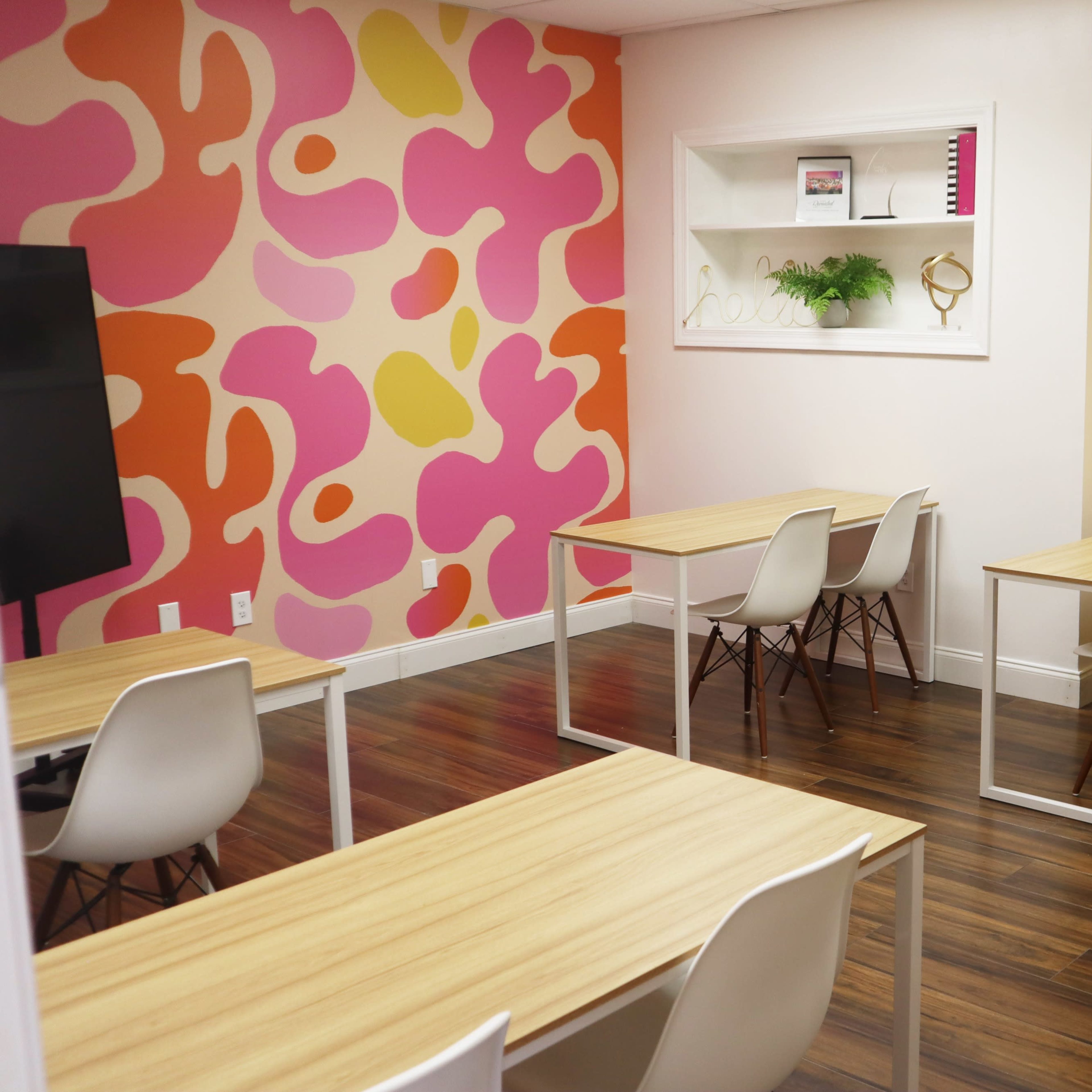 A brightly colored classroom with six wooden tables and chairs, set against a pink and yellow patterned wall.