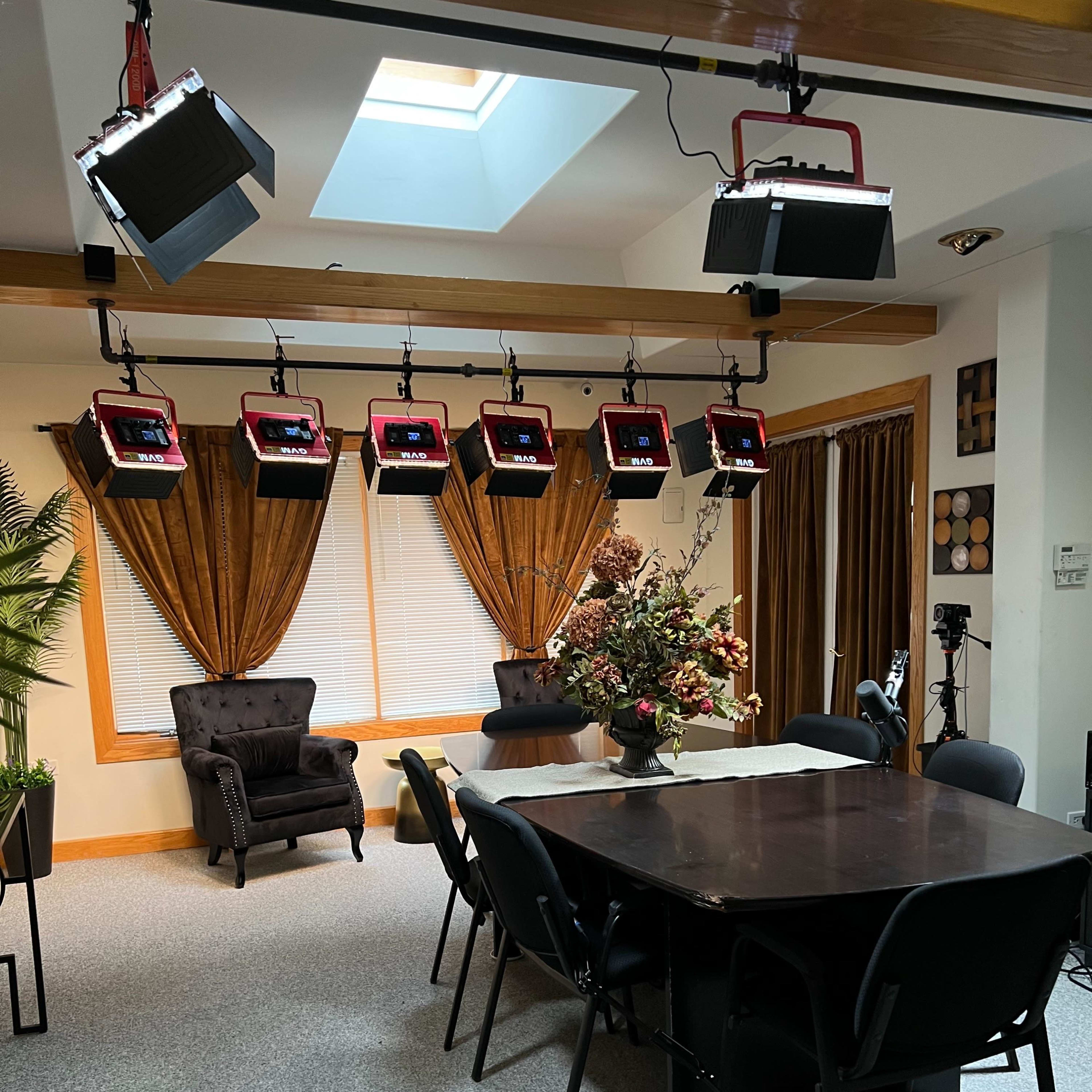 A well-lit room with a large table surrounded by chairs, decorated with a floral arrangement, and equipped with overhead lights and curtains.