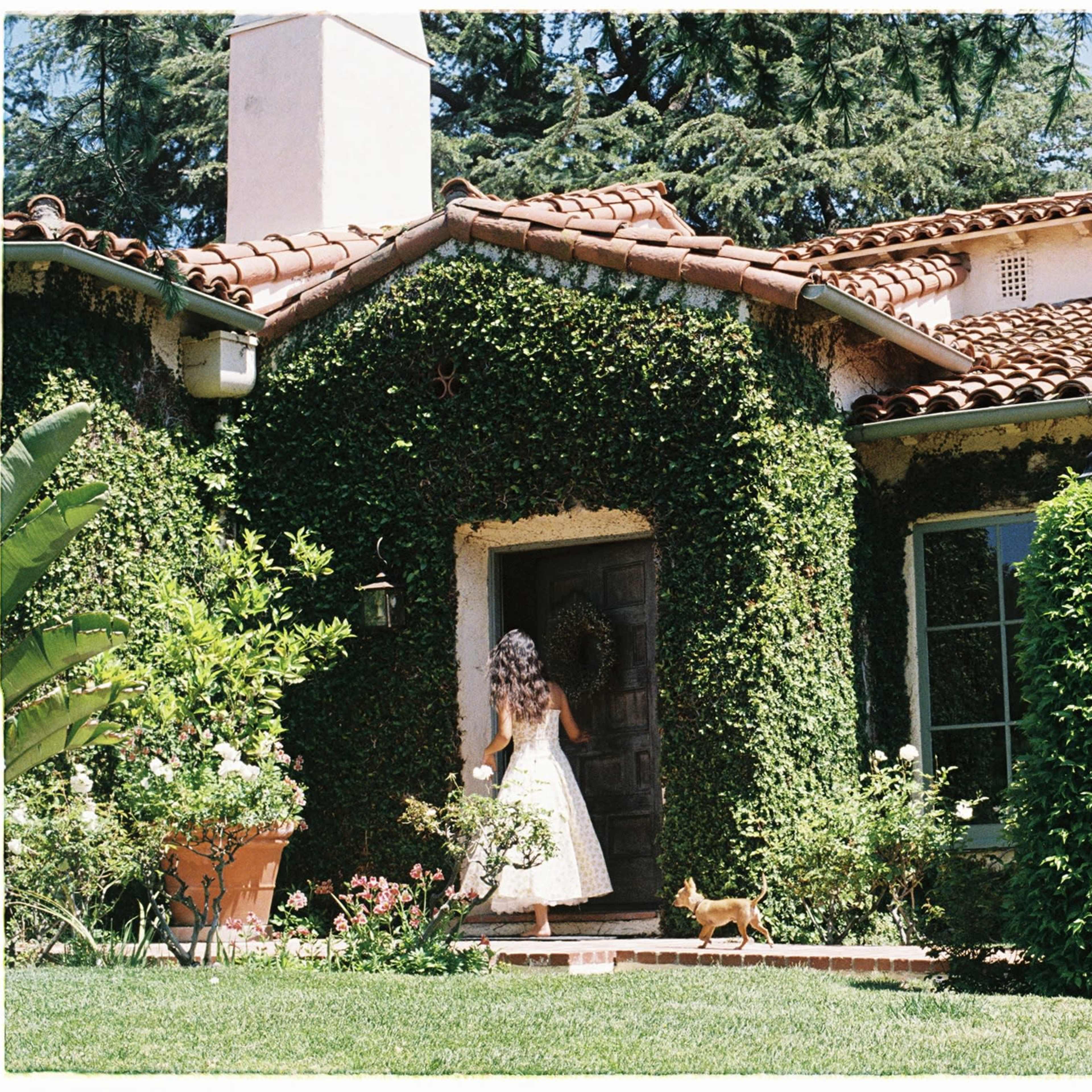 A girl wearing a white dress walks toward a house covered in ivy, with a small dog following her.