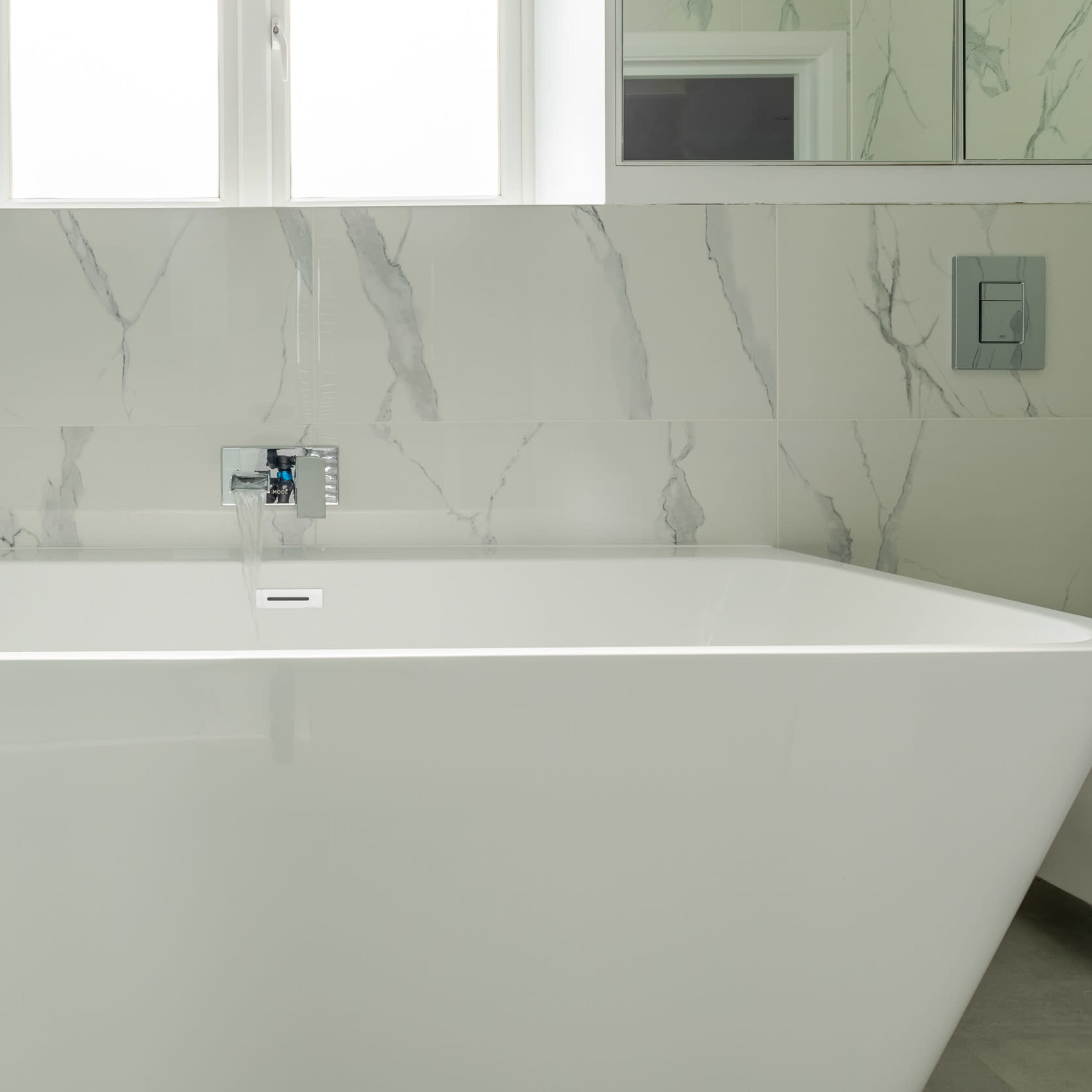 A modern, freestanding bathtub is positioned against a wall of marble tiles, with a window providing natural light above it.