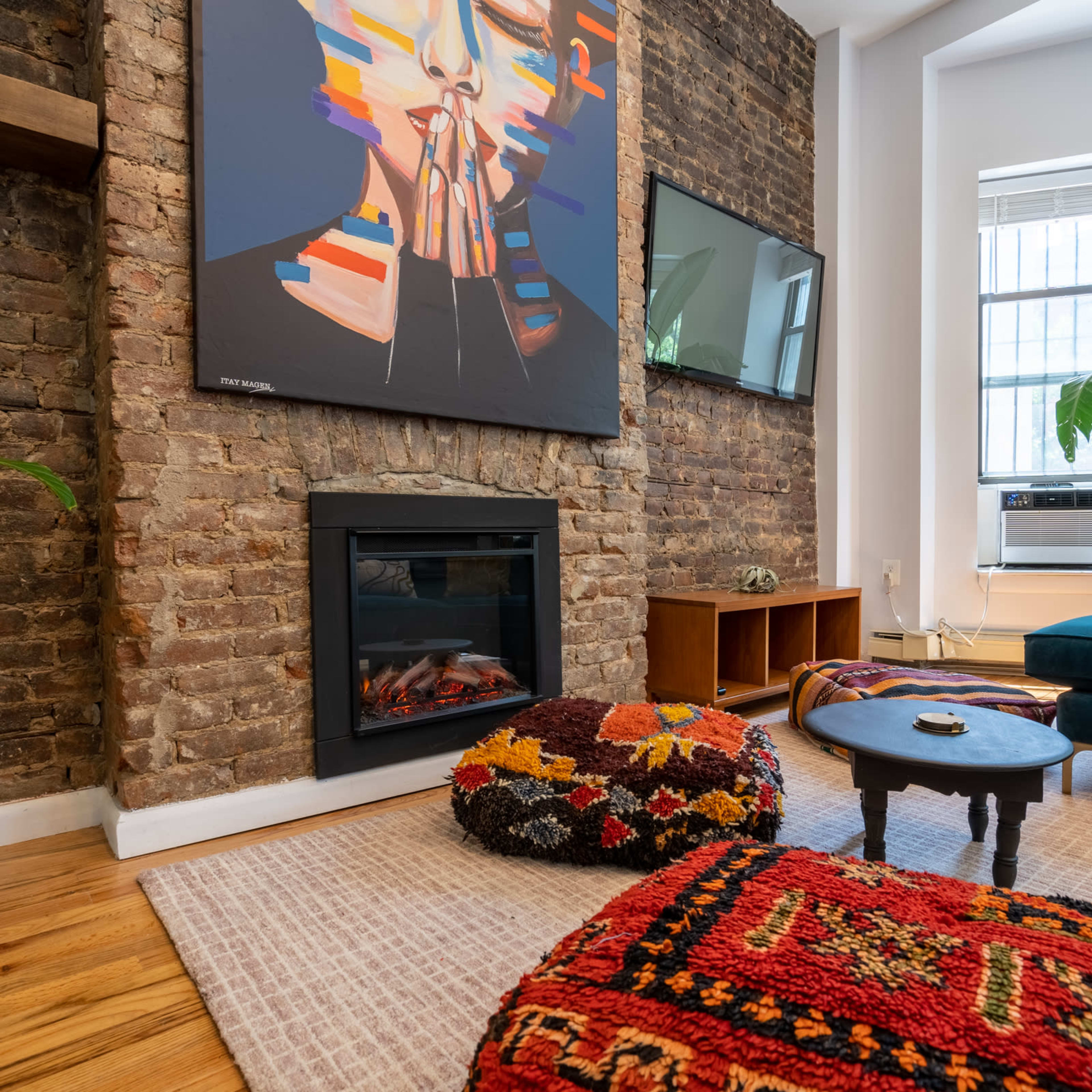A cozy living room with a brick wall, a modern electric fireplace, a colorful abstract painting, and a mix of patterned rugs with plants enhancing the decor.