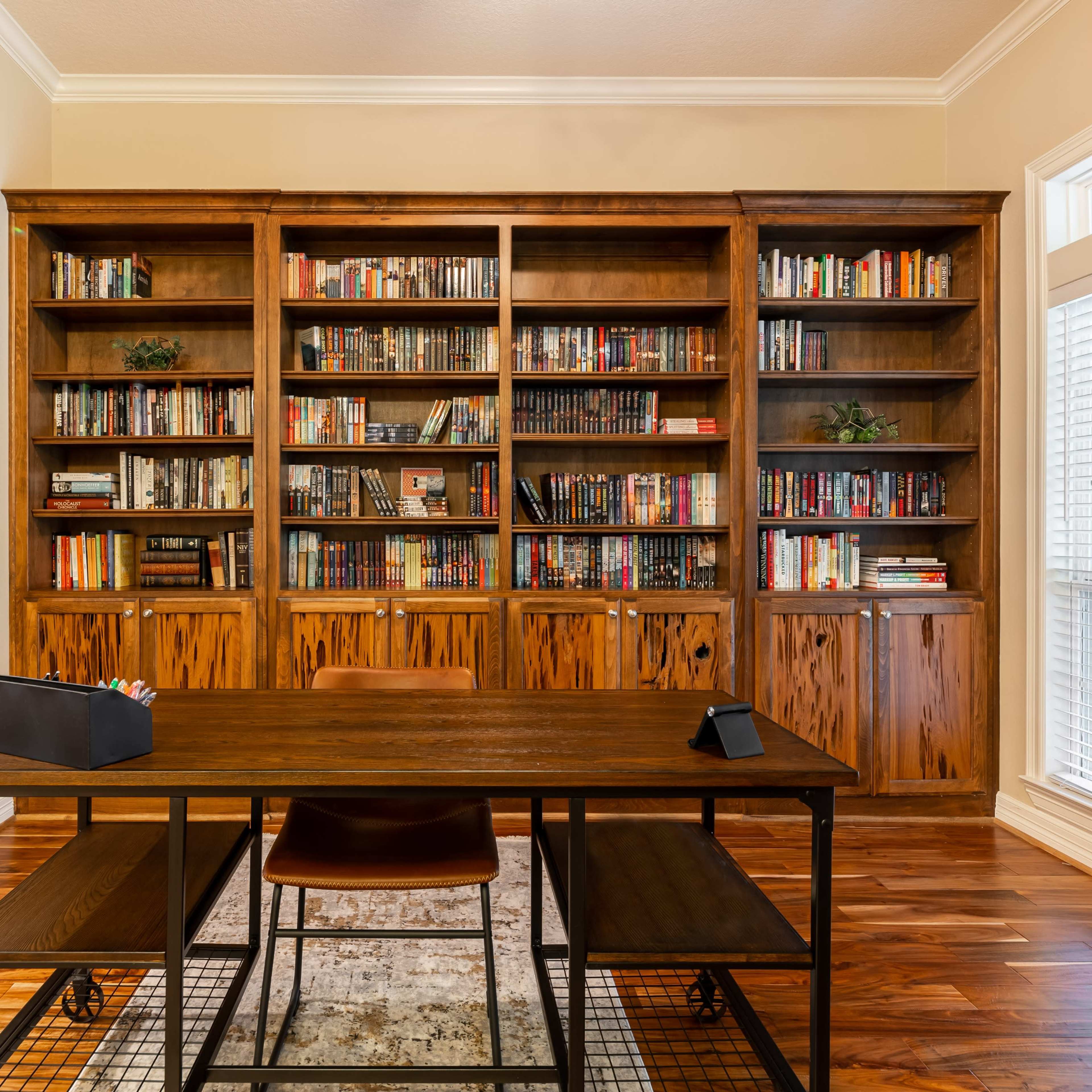 A wooden desk is positioned in front of a large bookshelf filled with books, accompanied by a potted plant in the corner of a well-lit room.