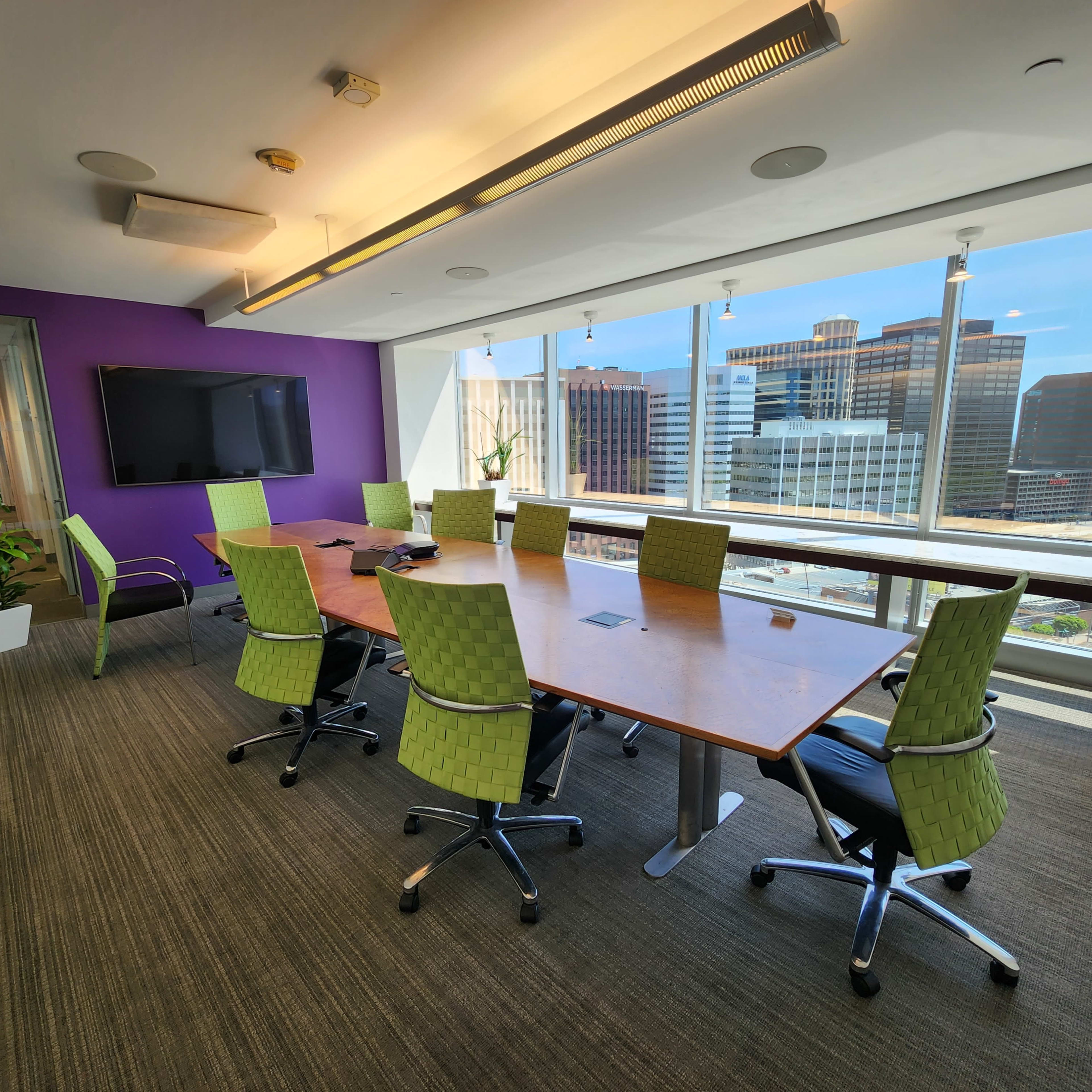 A modern conference room features a large wooden table surrounded by green ergonomic chairs, with a large window showcasing a city skyline.