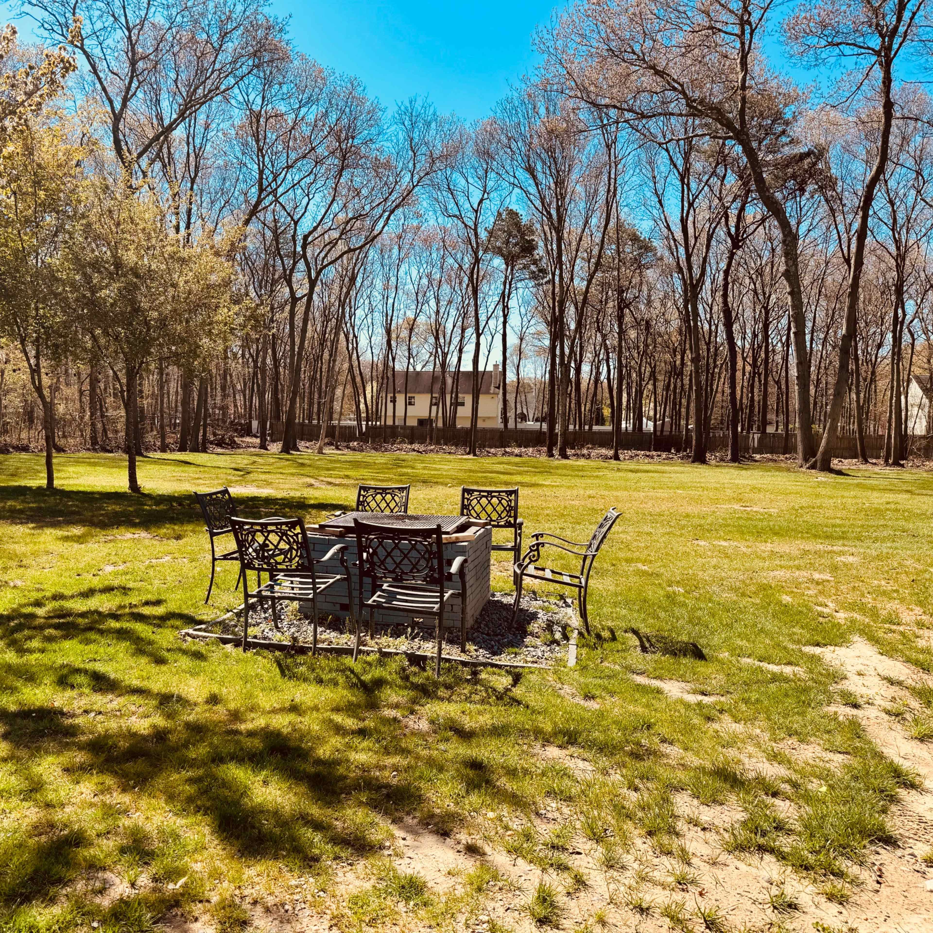A circular seating area with metal chairs surrounds a stone fire pit, set on a grassy lawn bordered by trees.