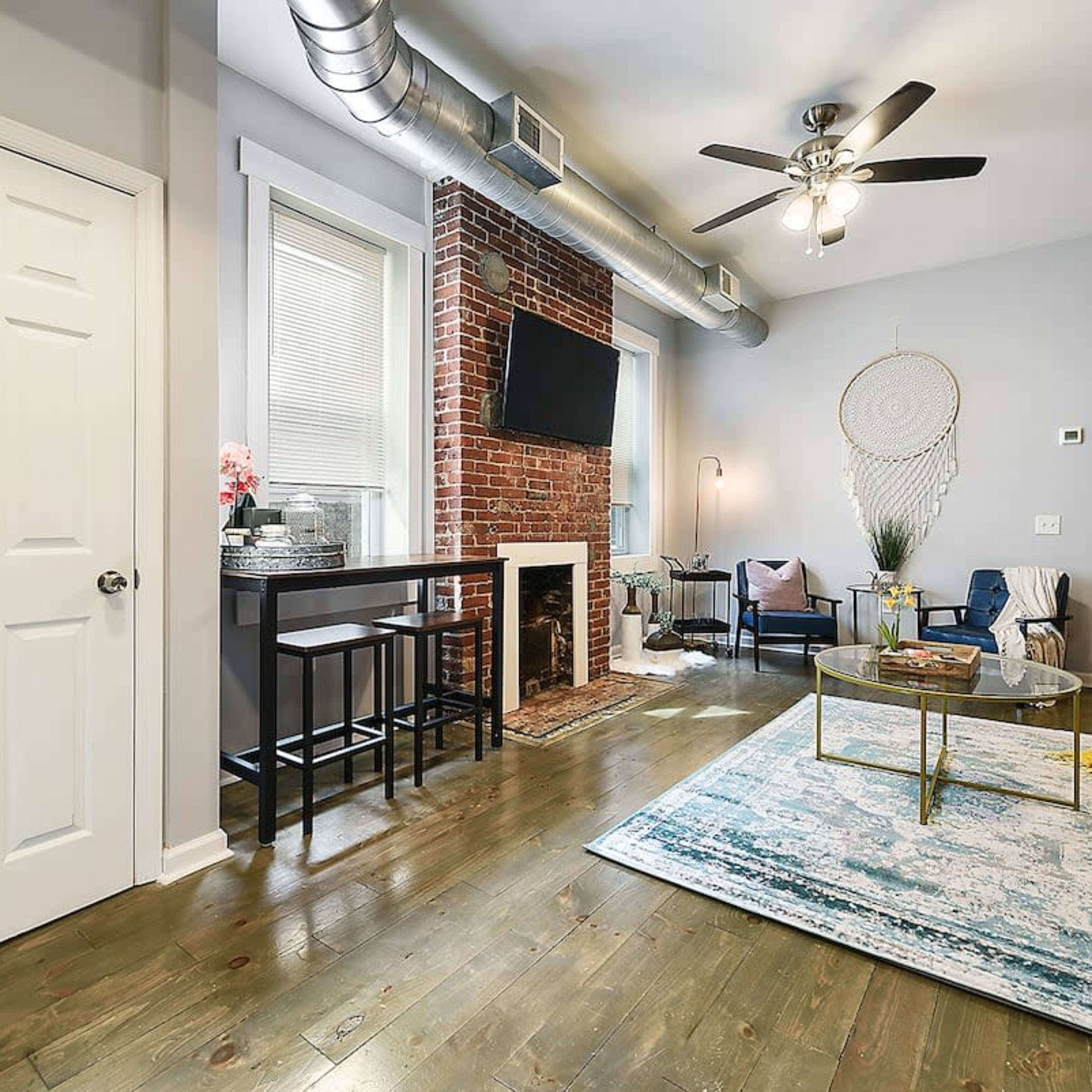 A modern living room featuring a sofa, coffee table, and a small dining area, with exposed brick walls and a ceiling fan.