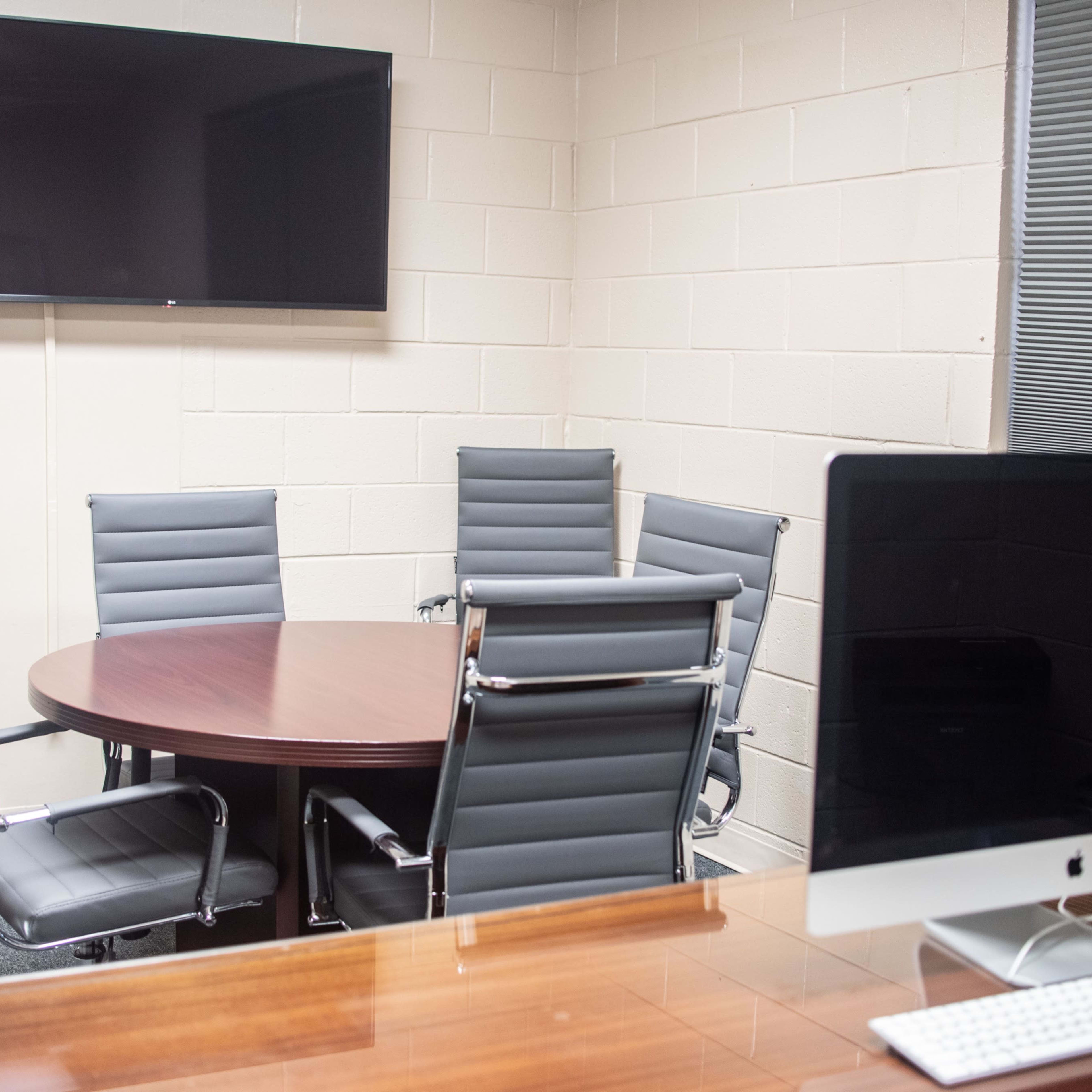 A conference room features a round table surrounded by five chairs, a wall-mounted television, and a computer on a desk.