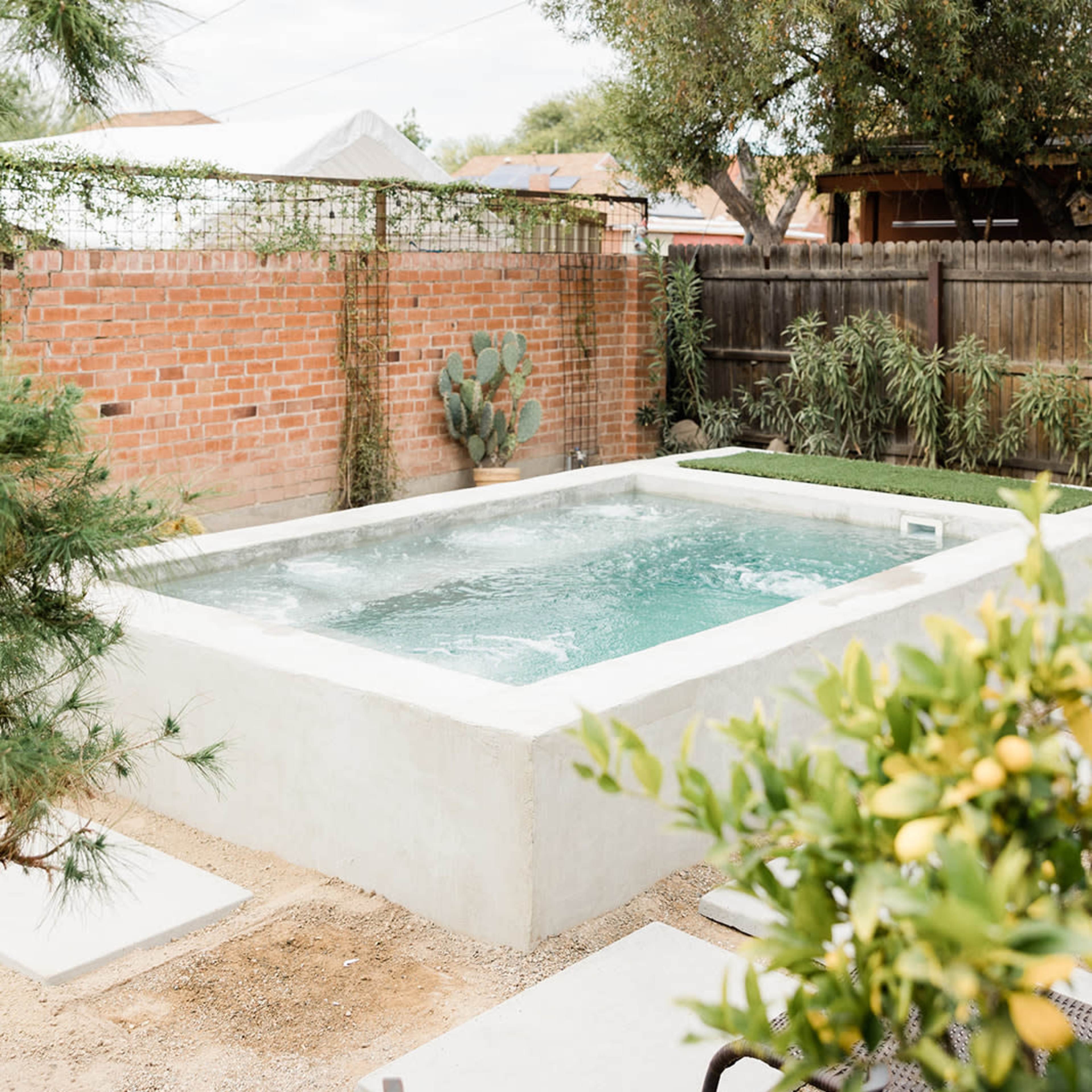 A rectangular pool with clear water is surrounded by a gravel area, adjacent to a brick wall and various plants.