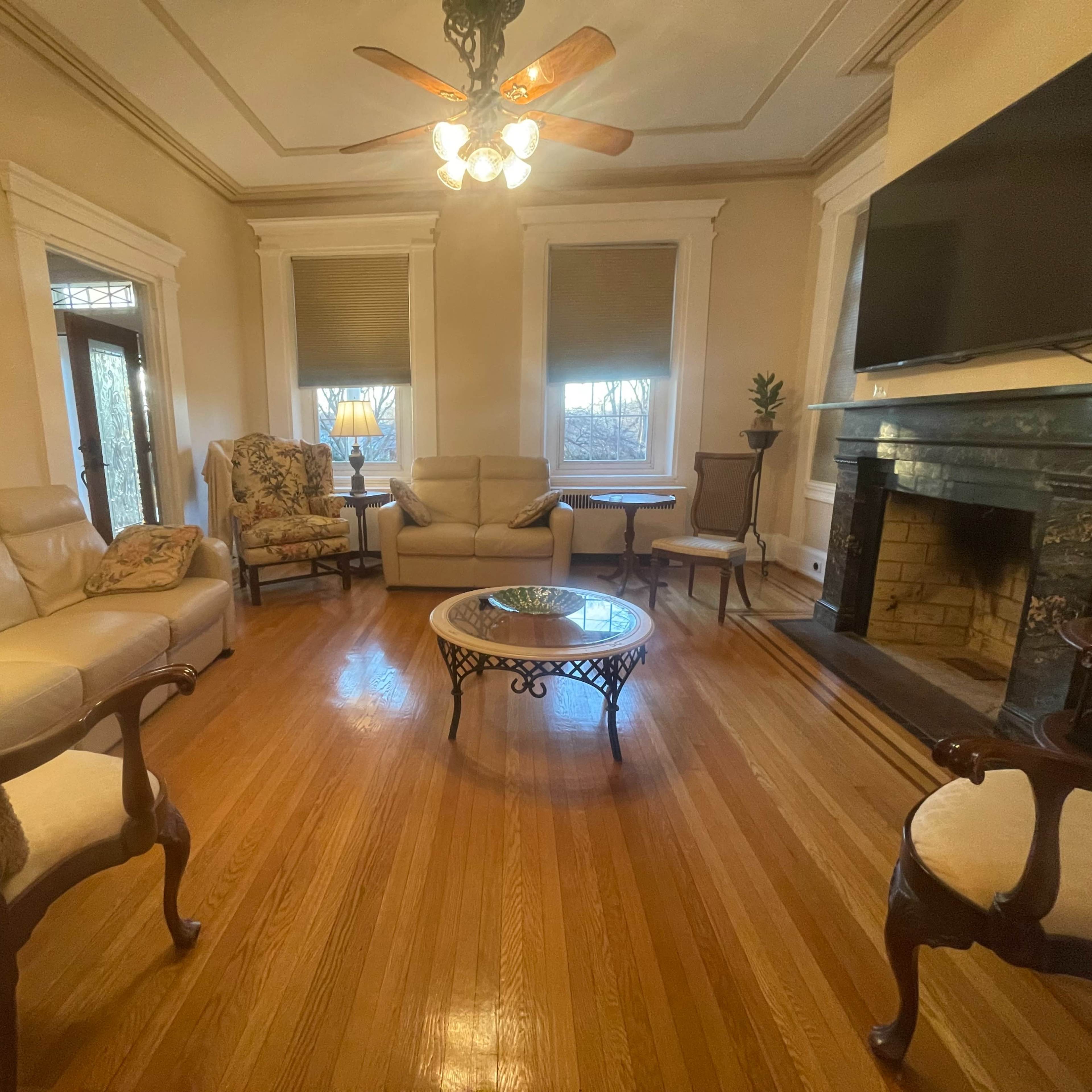 A traditional living room with hardwood floors, a seating arrangement including cream-colored sofas, a patterned armchair, and a coffee table, along with a fireplace and a large television mounted on the wall.