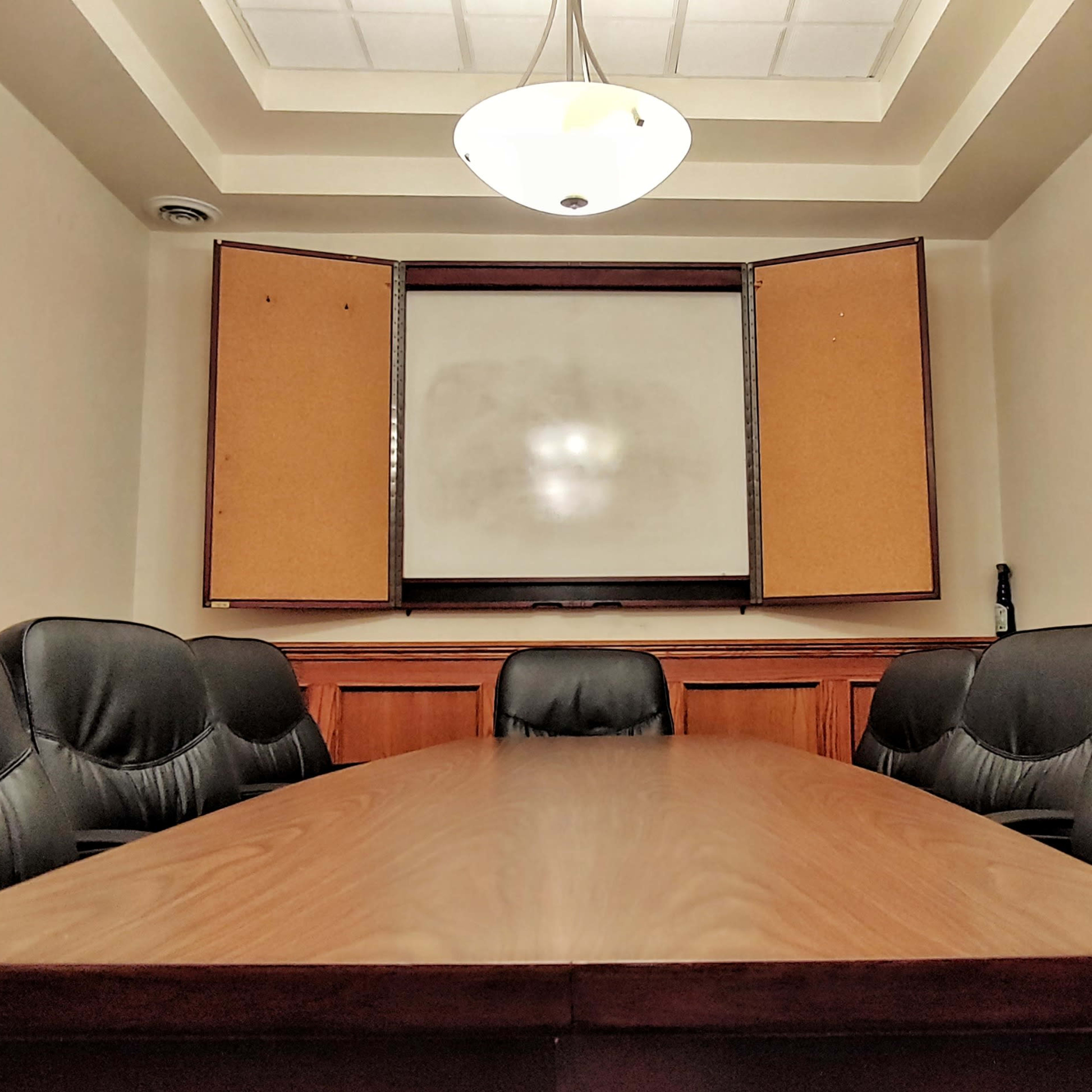 A conference room features a long wooden table surrounded by black leather chairs, with a whiteboard at one end and windows partially open.