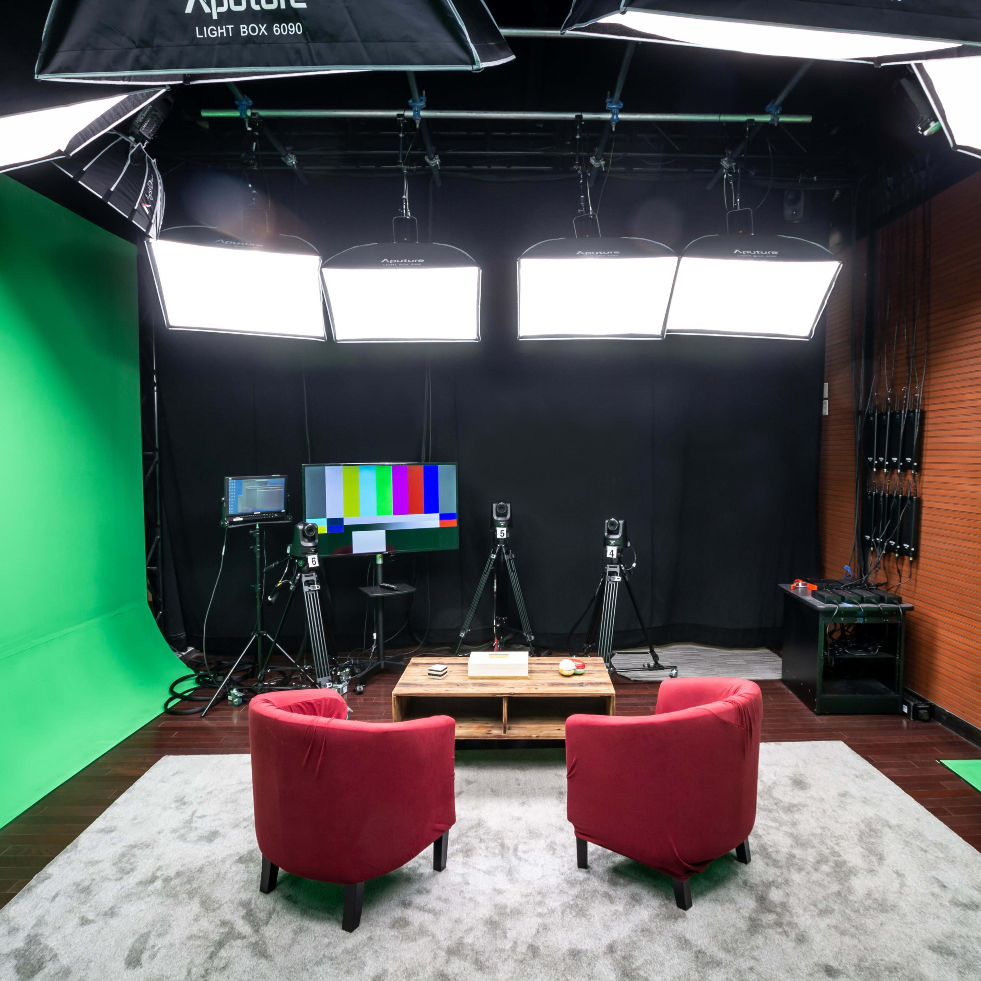 A video recording studio with two red chairs facing a wooden table, surrounded by green and black backdrop panels and equipped with multiple lights and cameras.