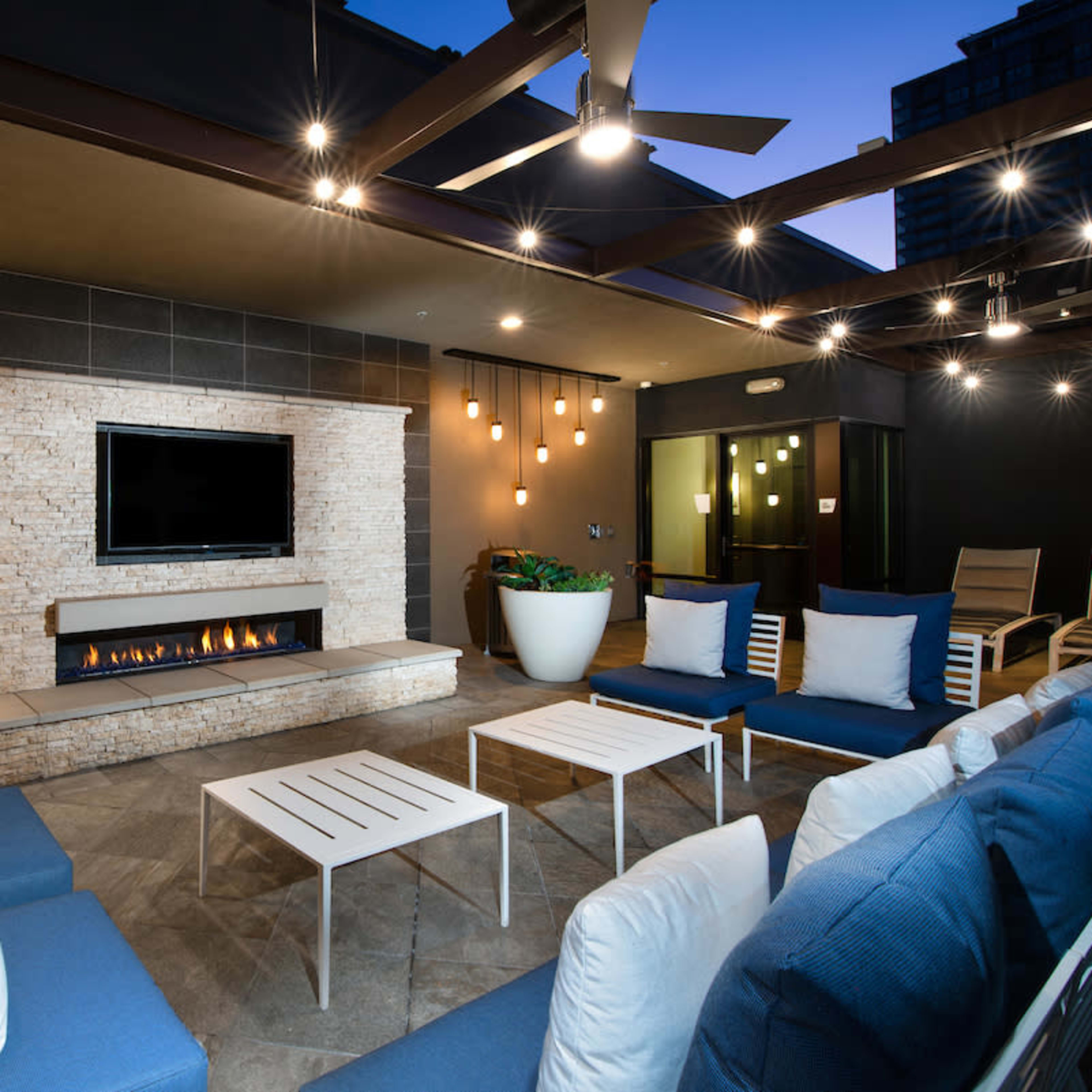 A modern outdoor lounge area features a stone fireplace, a flat-screen TV, blue seating, and decorative lighting.
