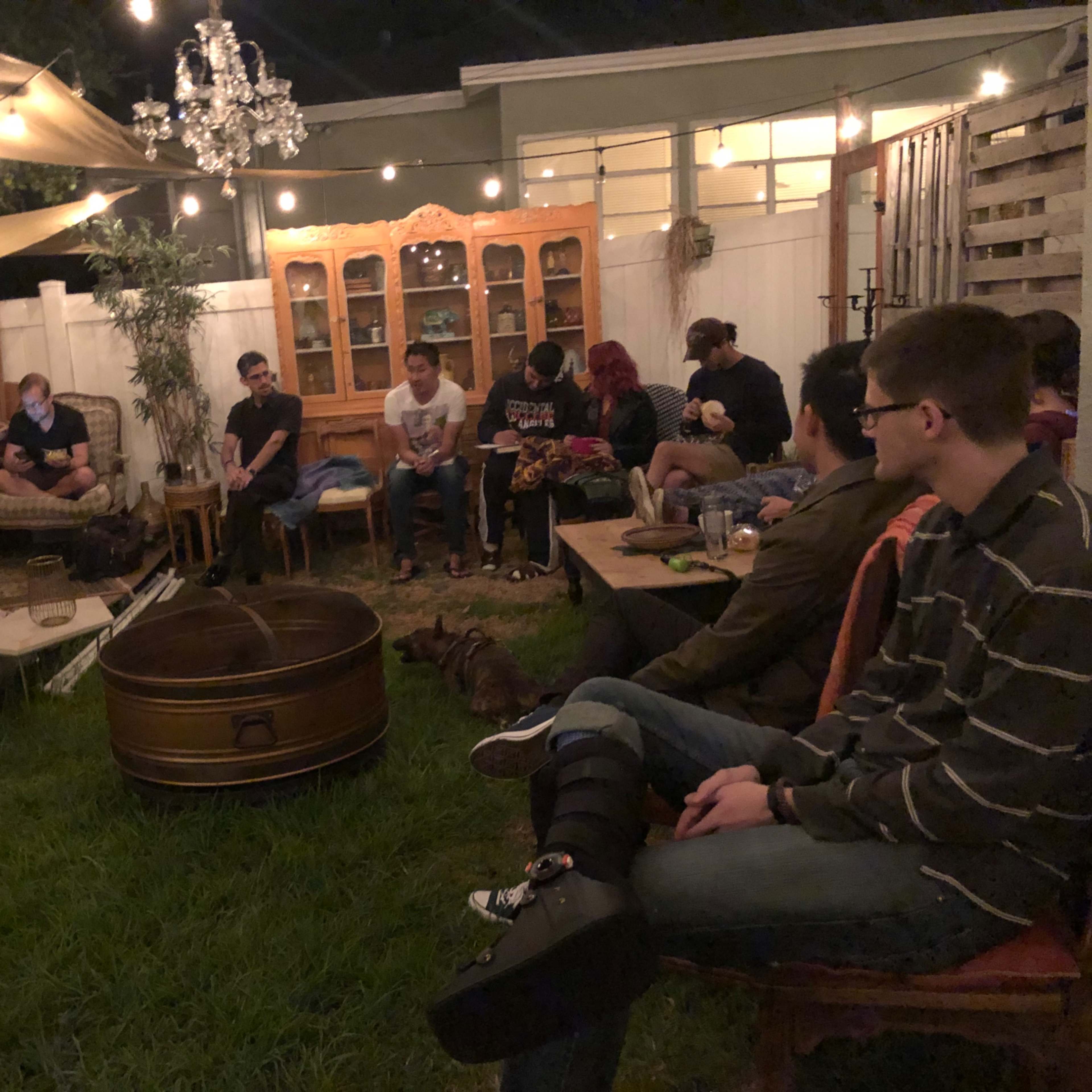 A group of people sits in a well-lit backyard, arranged in a circle around a central fire pit, with various seating options and a decorative cabinet in the background.