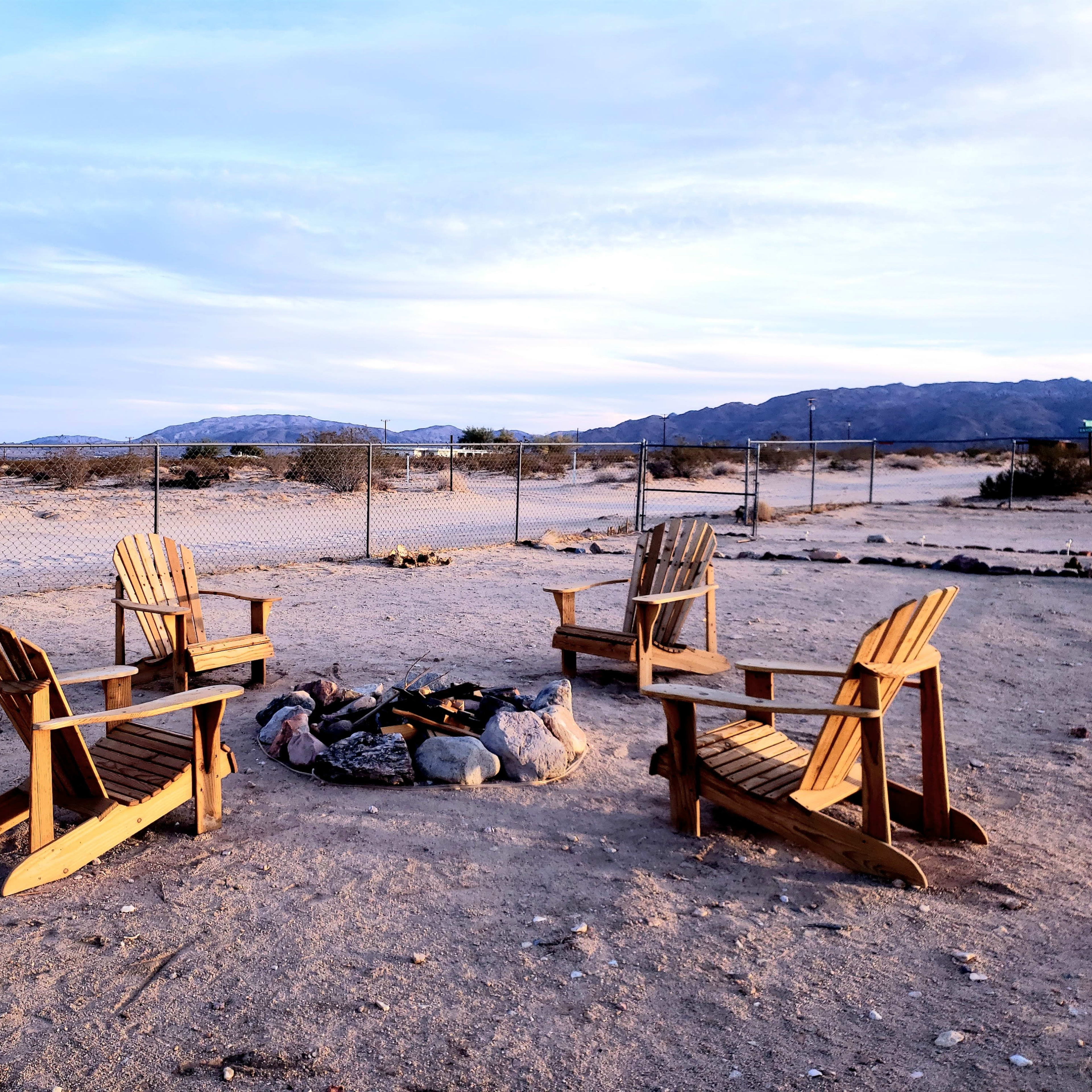 Four wooden chairs are arranged in a circle around a fire pit in a sandy outdoor area surrounded by a fence and desert landscape.