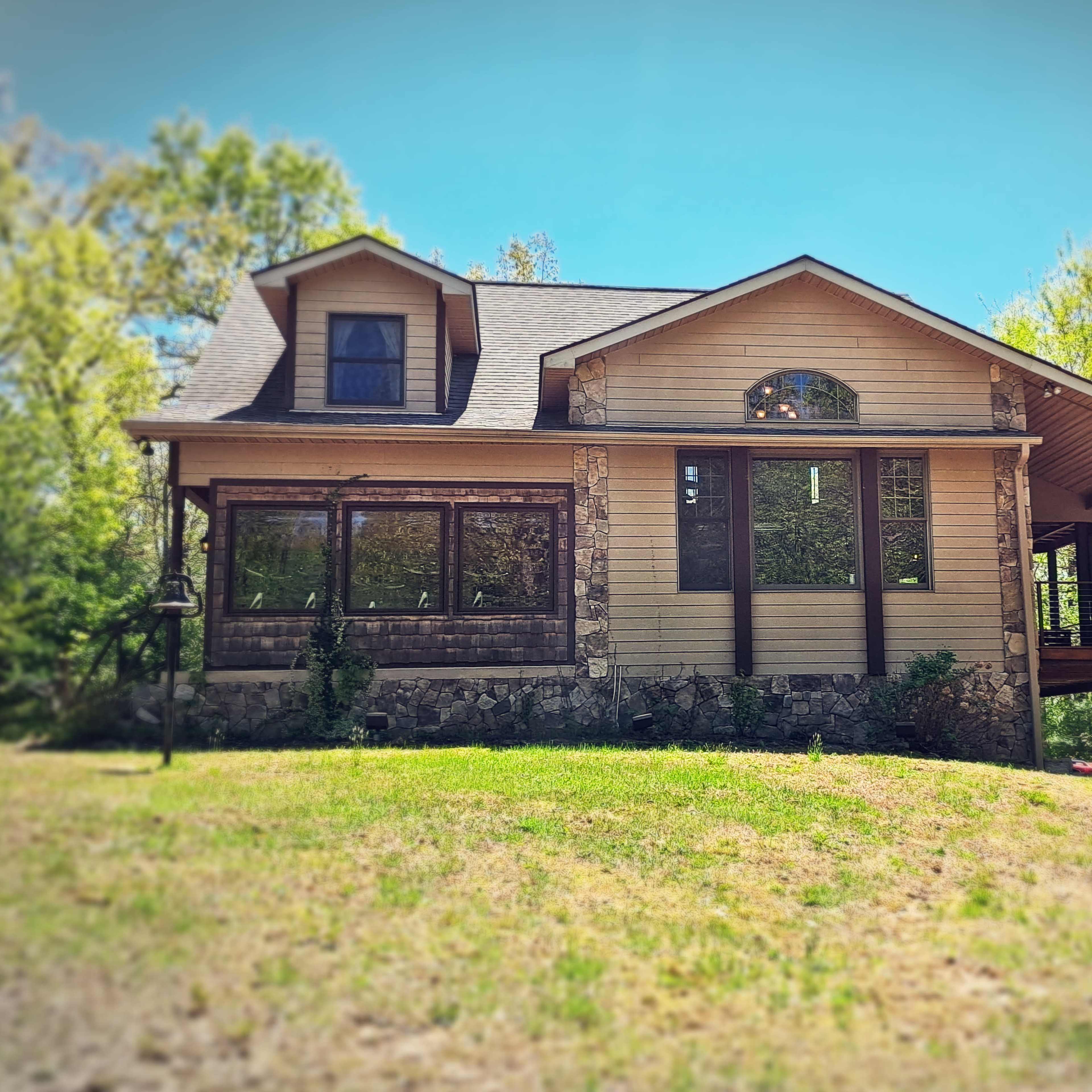 A two-story house with a stone foundation and large windows is surrounded by green trees.