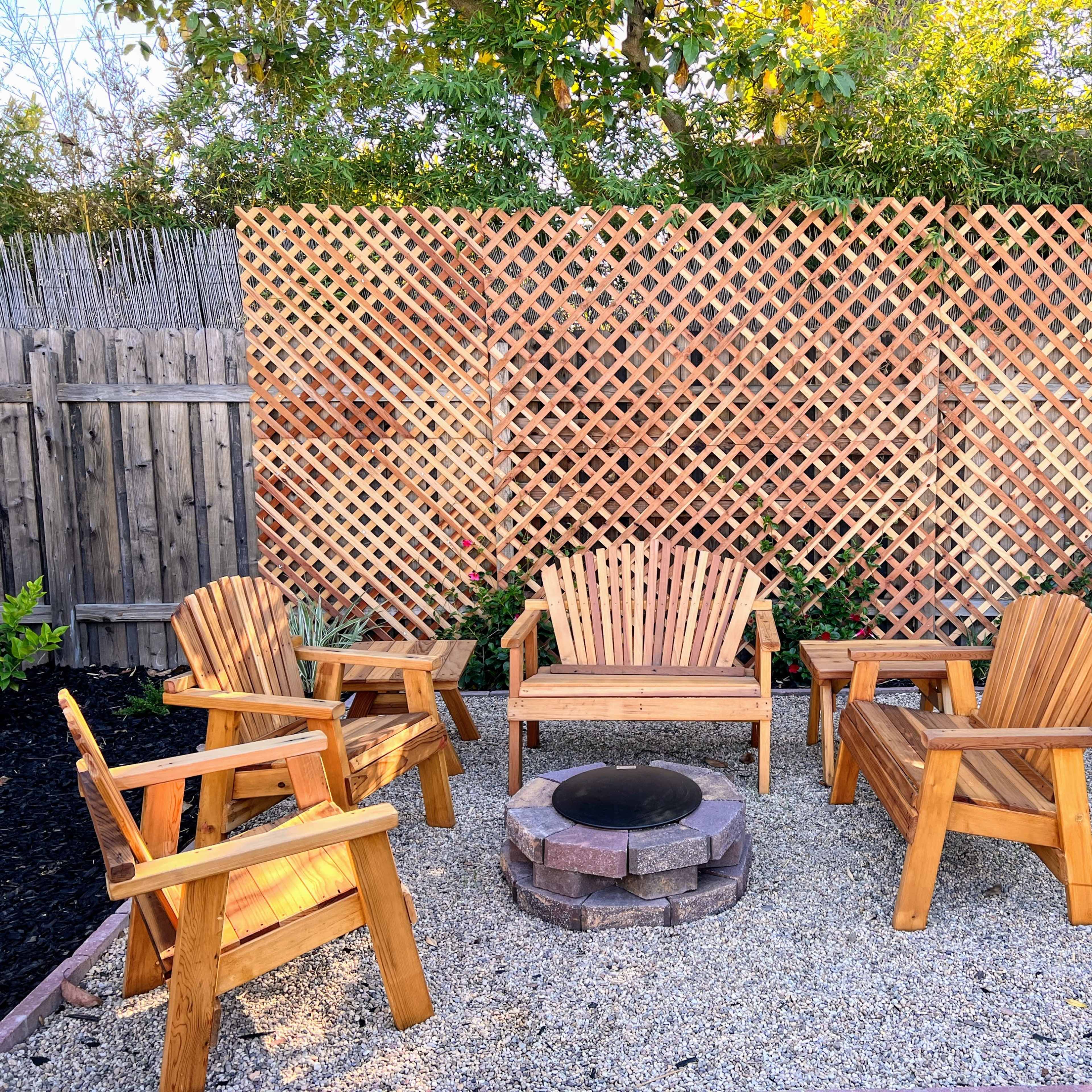 A gravel patio with wooden chairs and a bench arranged around a circular stone fire pit, set against a backdrop of a wooden lattice fence and green plants.