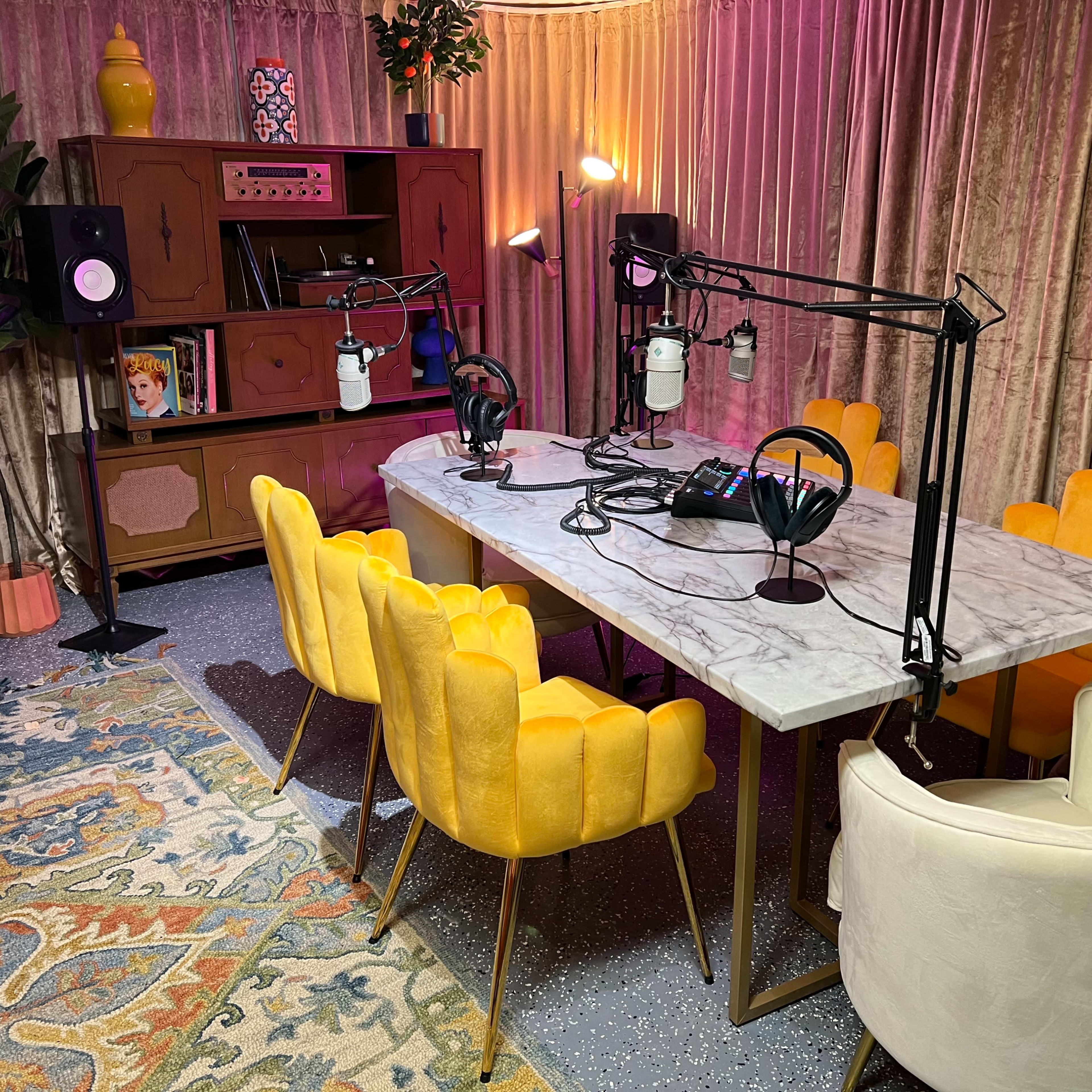 A podcast recording studio featuring a marble table surrounded by yellow and white chairs, with microphones mounted on stands and a retro-style cabinet in the background.