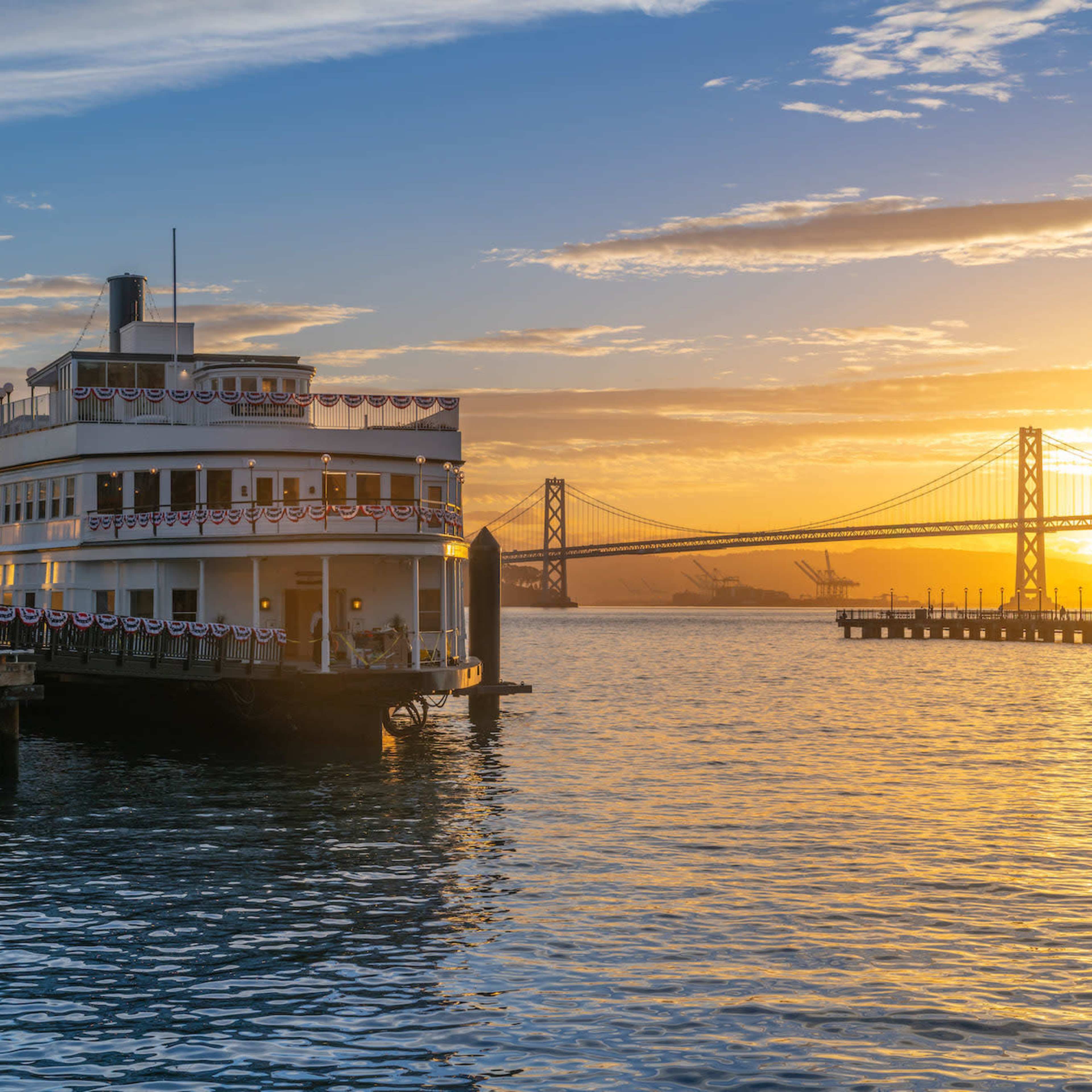 A large boat adorned with decorations is docked at a pier during sunset, with the Bay Bridge visible in the background.